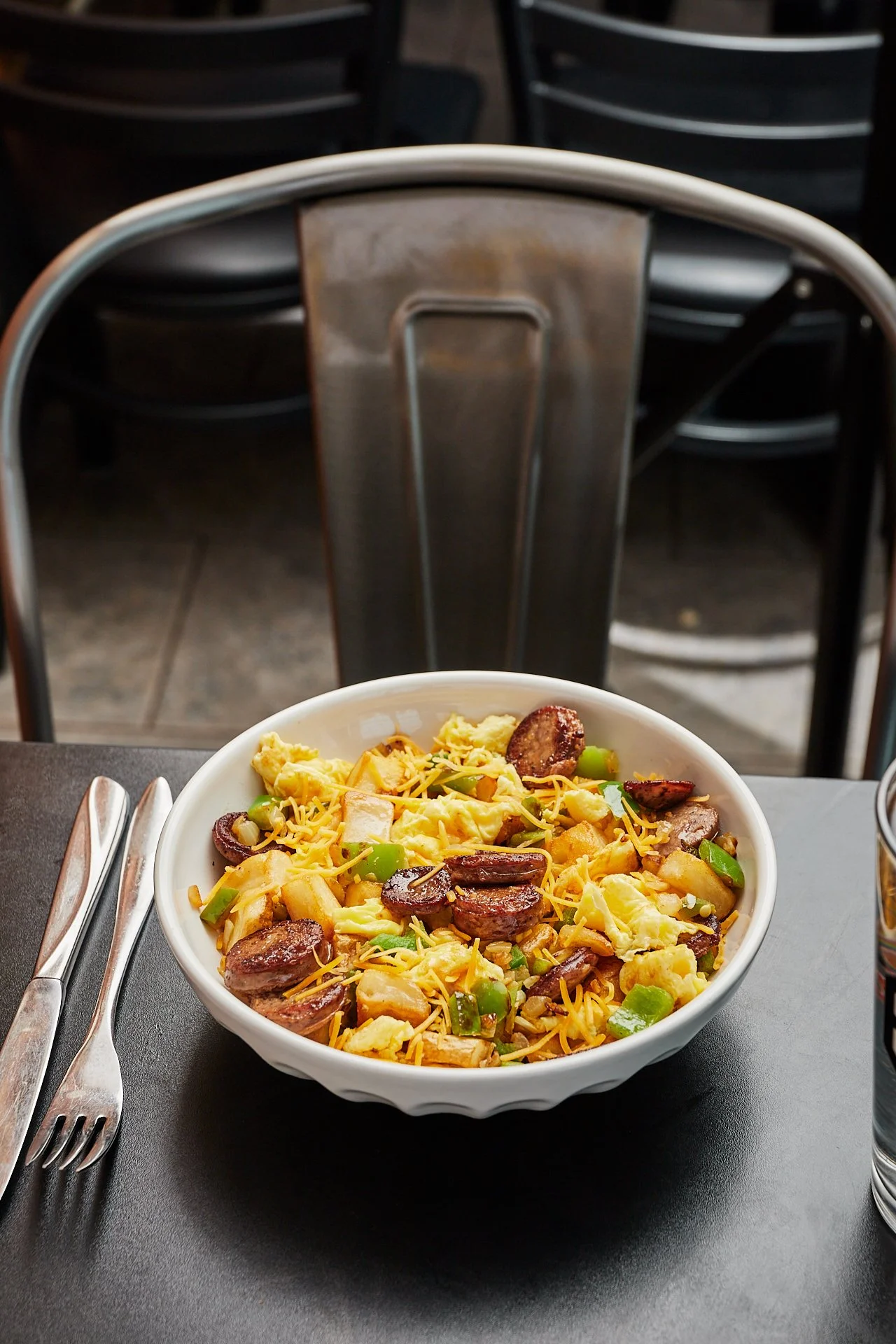 A bowl of breakfast scramble with eggs, sausage slices, green bell peppers, and shredded cheese on a black table, with a fork and knife beside it, in a restaurant setting.