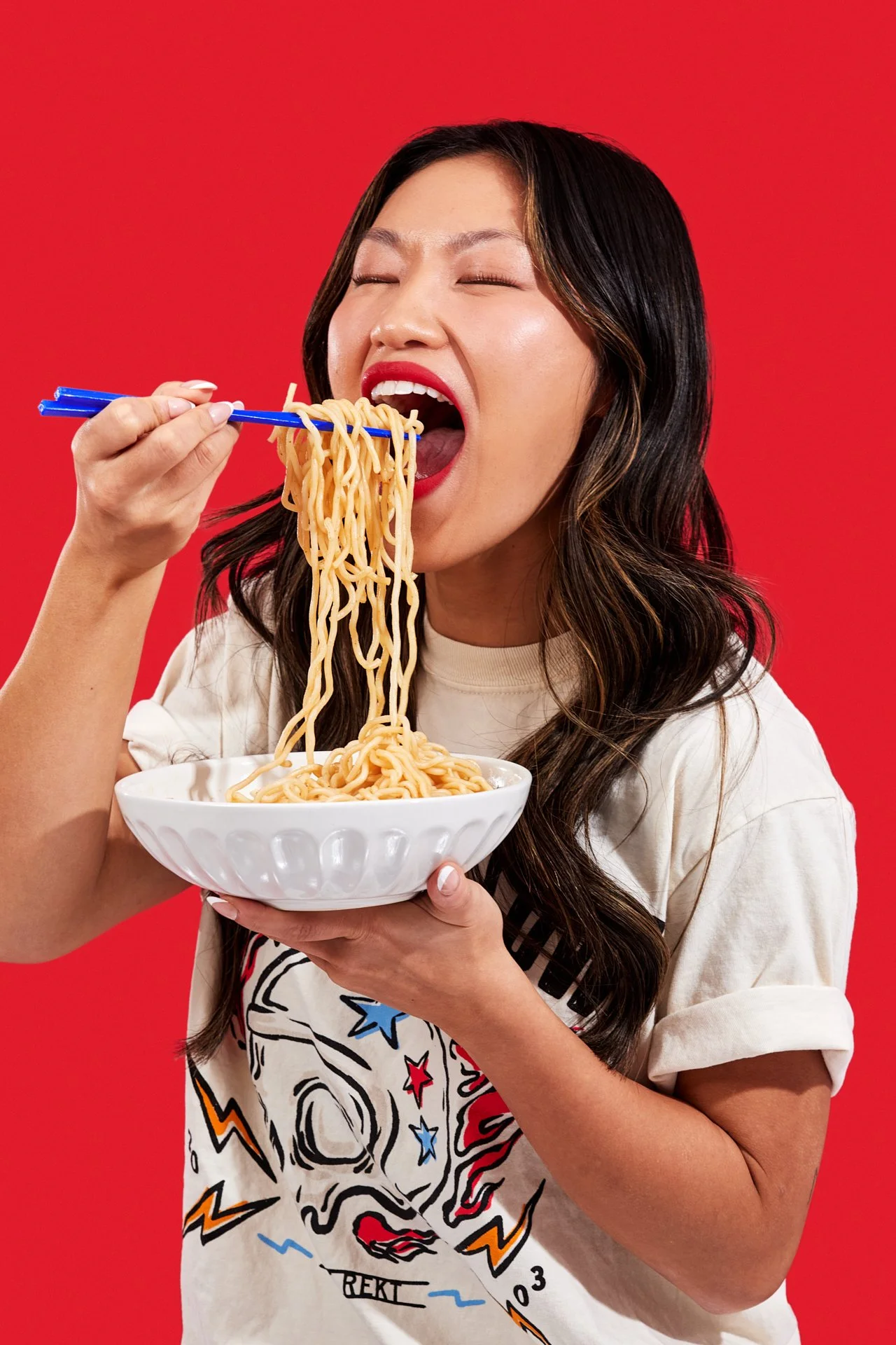 A woman with dark hair and red lipstick is holding a bowl of ramen noodles and eating with blue chopsticks against a red background.