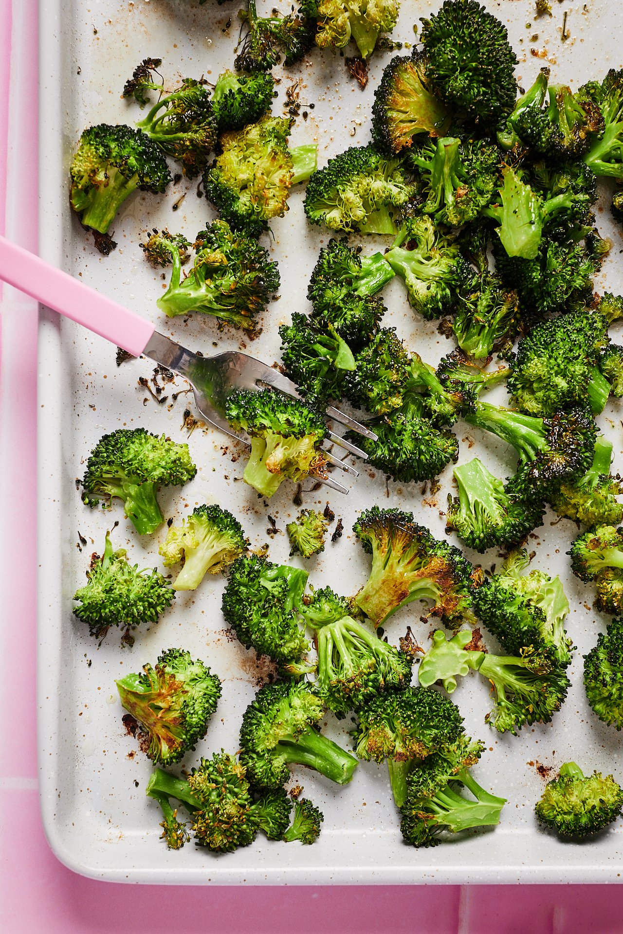 Roasted broccoli florets on a white baking sheet with a pink background, some florets slightly charred.