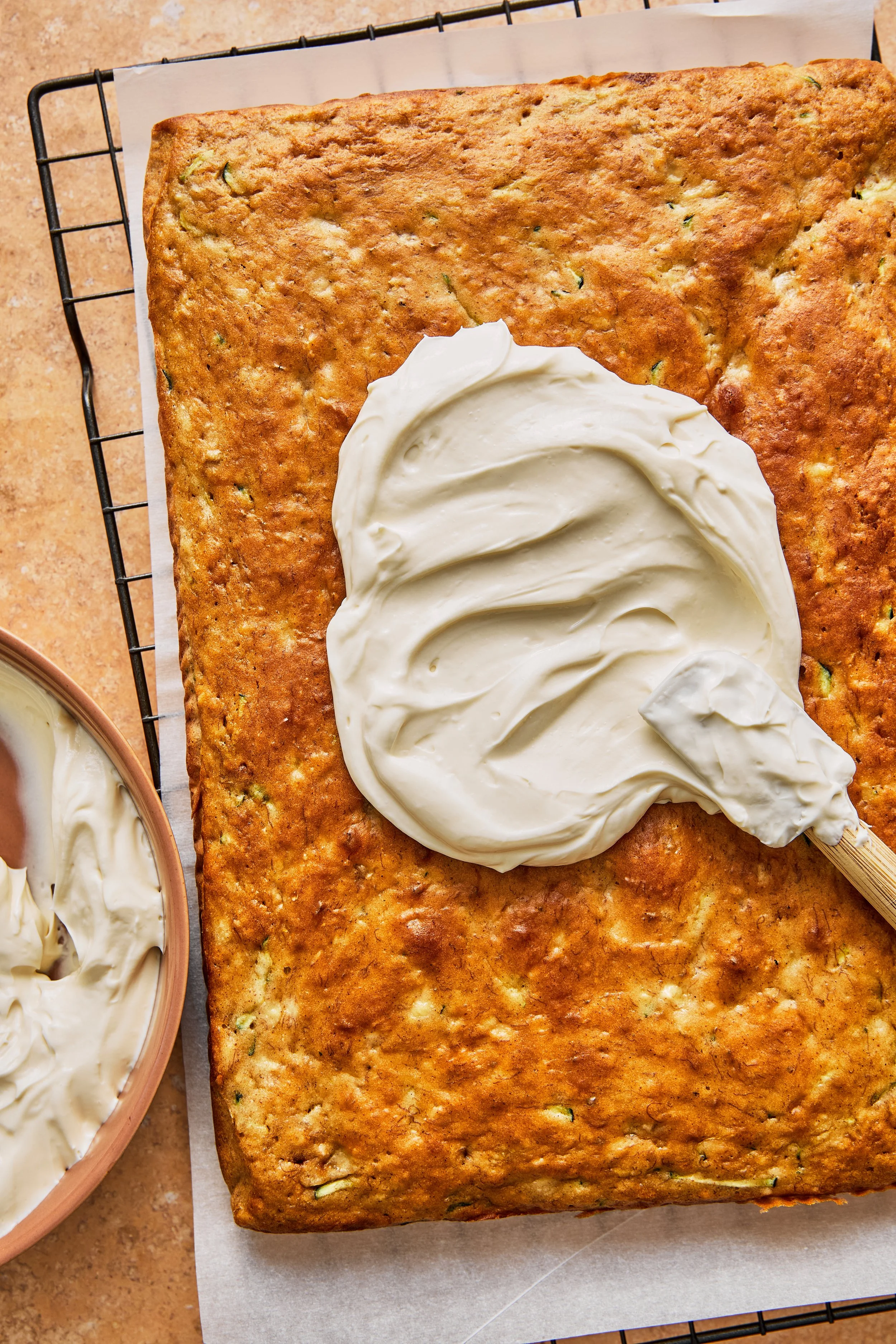 A large square zucchini loaf with a dollop of cream cheese frosting on top, cooling on a wire rack.