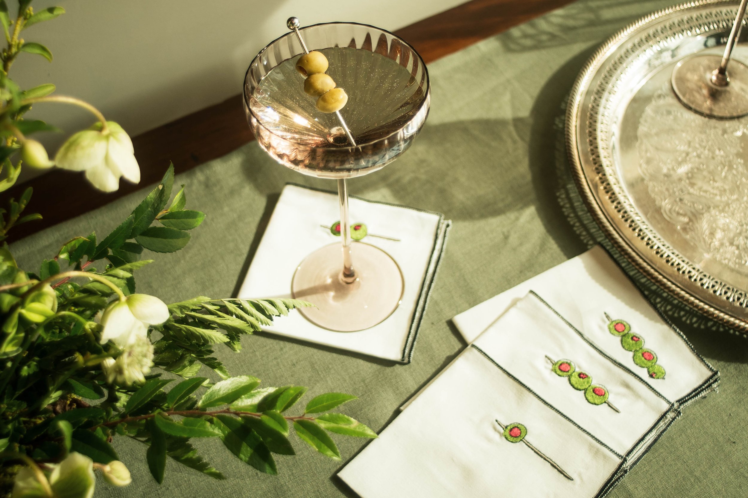 Olive Cocktail Napkins laid out on a stone green table runner next to a silver serving plate and a floral arrangement