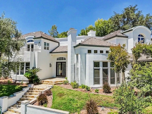 A large white house with multiple levels, rounded windows, and a tiled roof, surrounded by trees and landscaped yard with stone steps.