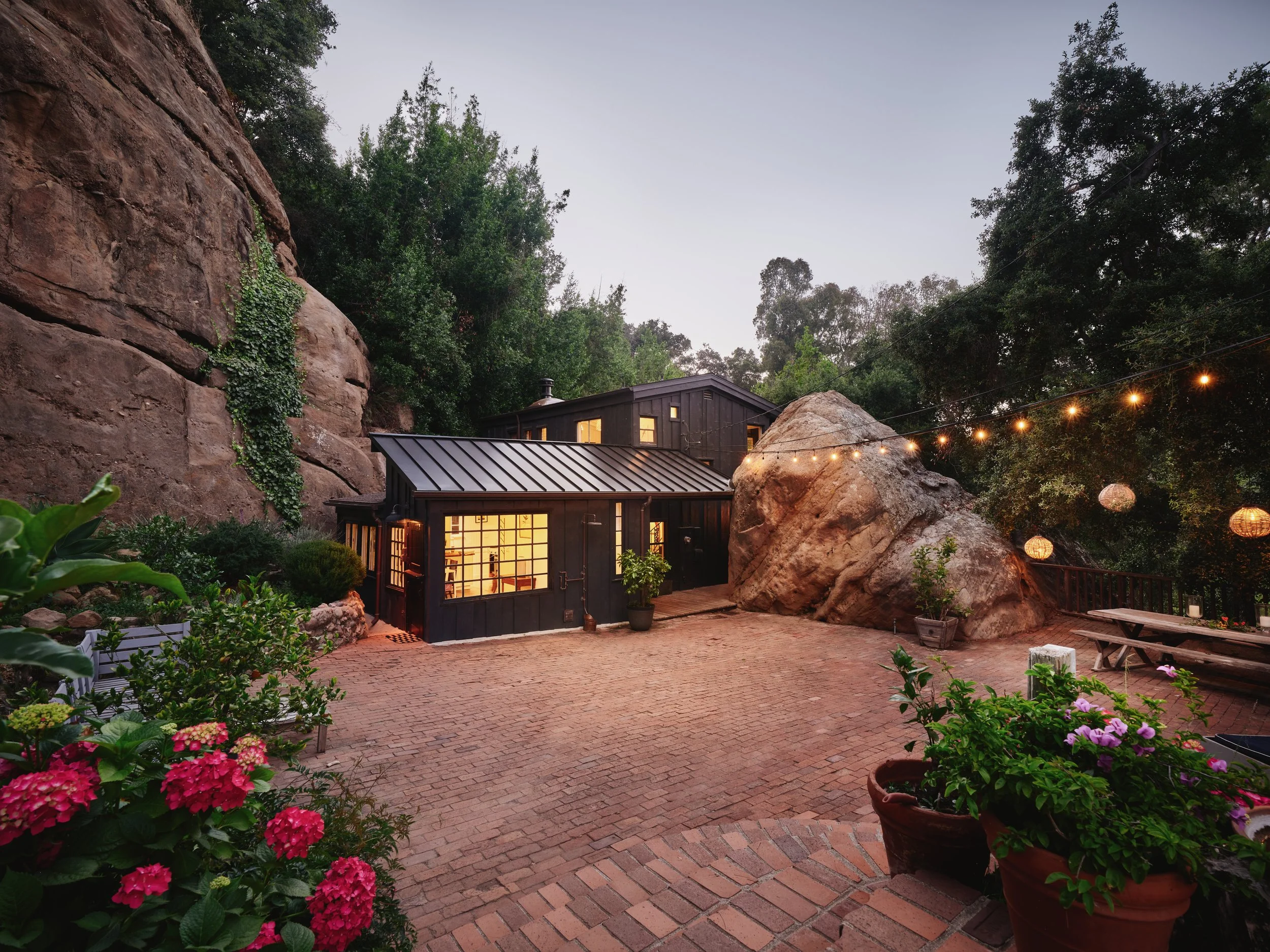 Cozy backyard with a rustic house, brick patio, and large rock formations surrounded by trees; string lights and potted plants enhance the ambiance.