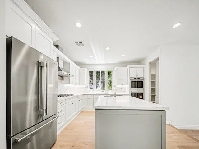 Bright, modern kitchen with white cabinetry, stainless steel refrigerator, and large island in the center, with a window and built-in appliances.