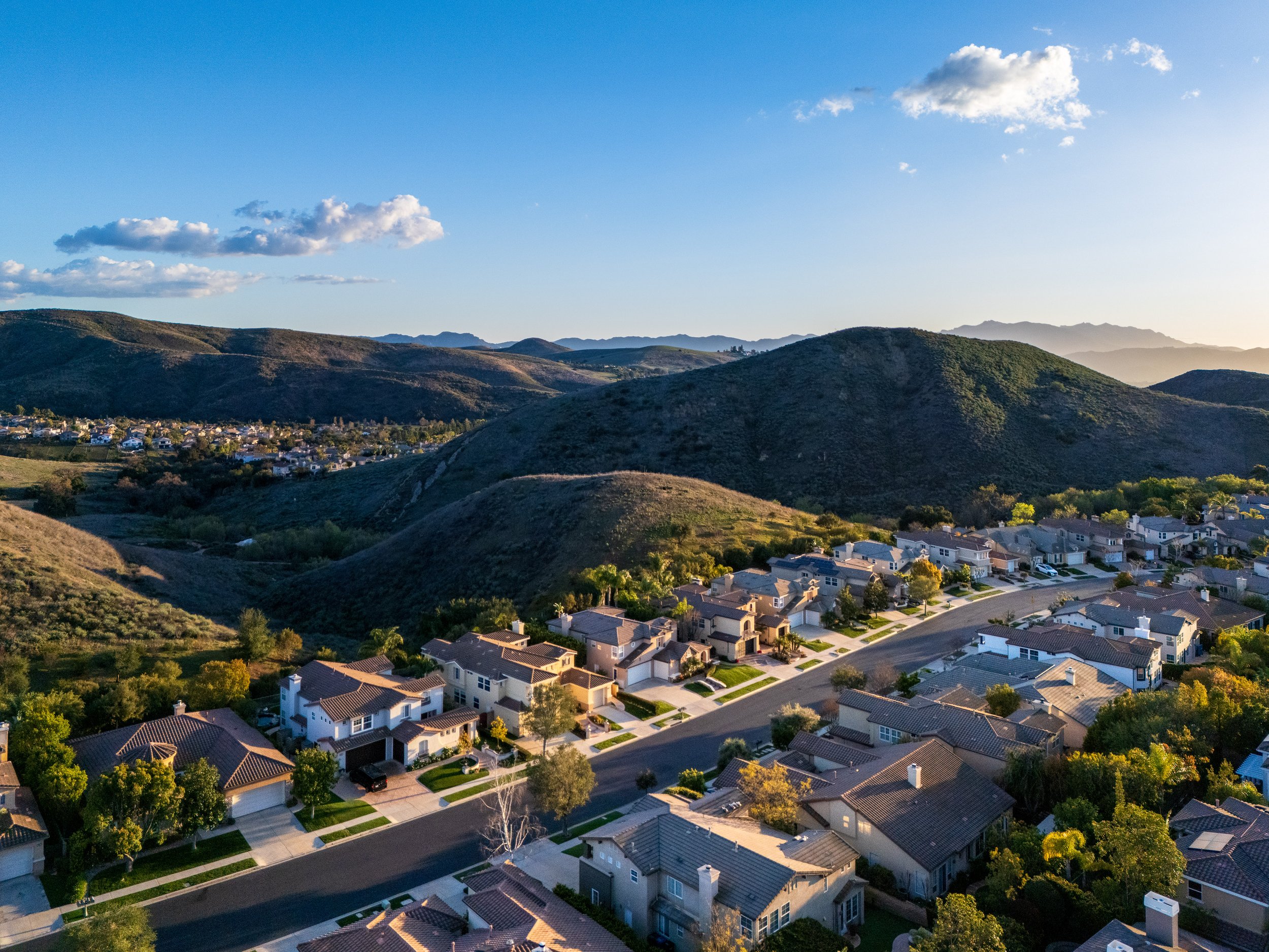 Aerial view of a residential neighborhood with houses and streets, surrounded by green hills under a clear blue sky with a few clouds.