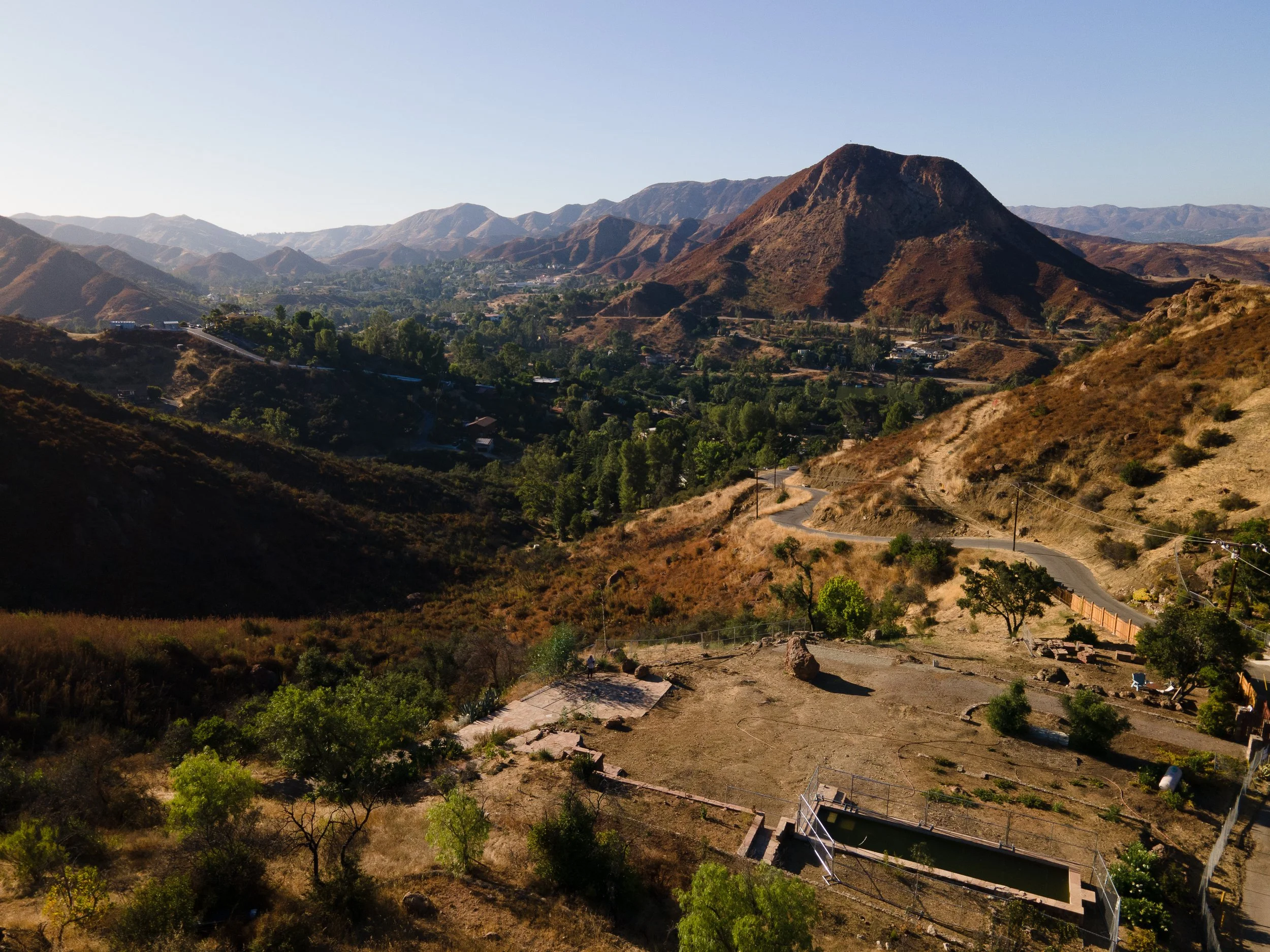 Panoramic view of a hilly landscape with a prominent mountain, surrounded by green trees and vegetation. A winding road and scattered buildings are visible, with a dry, brown terrain in the foreground under a clear blue sky.