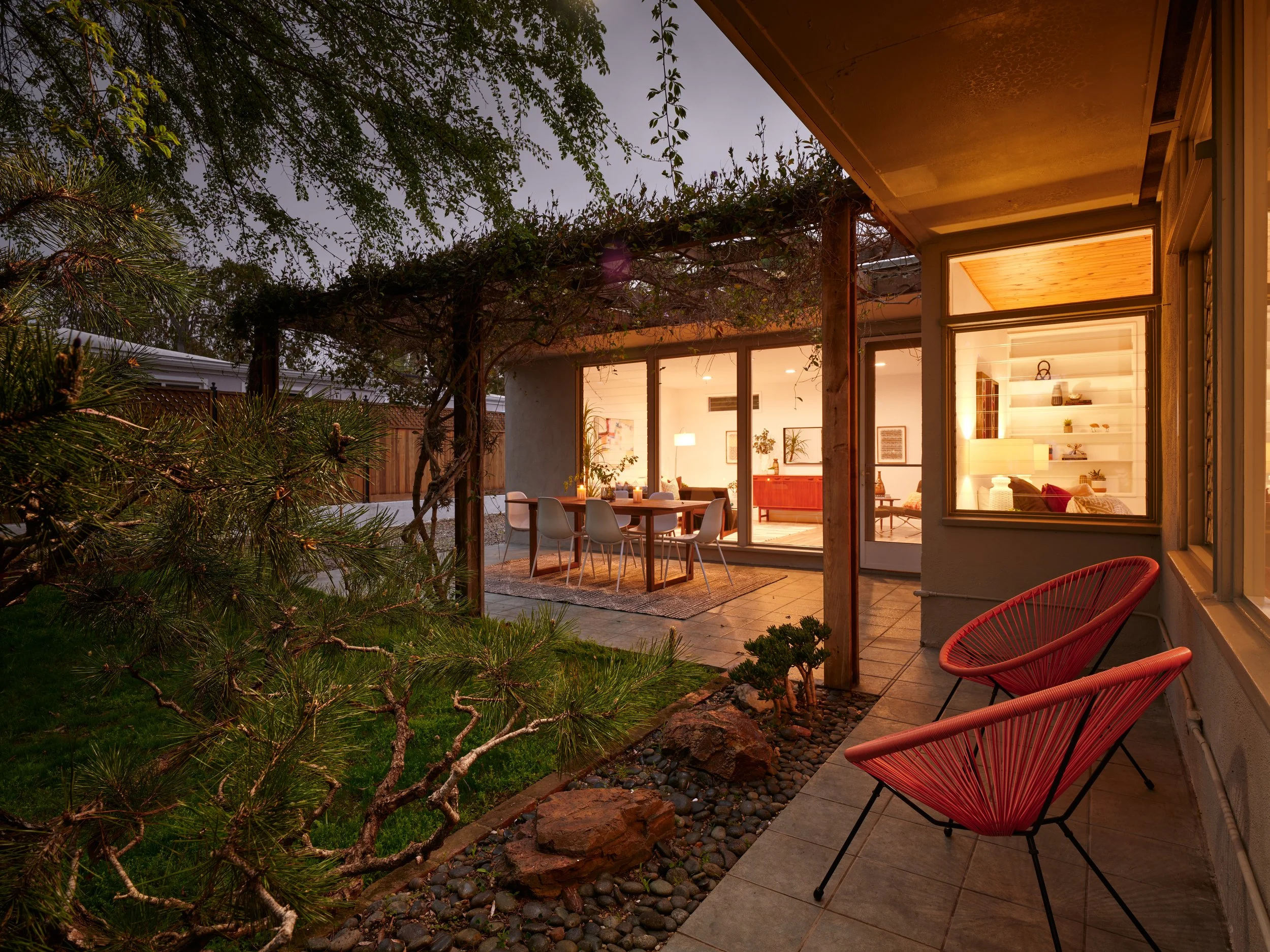 Outdoor patio with red chairs and garden view, leading to a modern interior with illuminated dining area and living space with shelves, visible through glass doors.