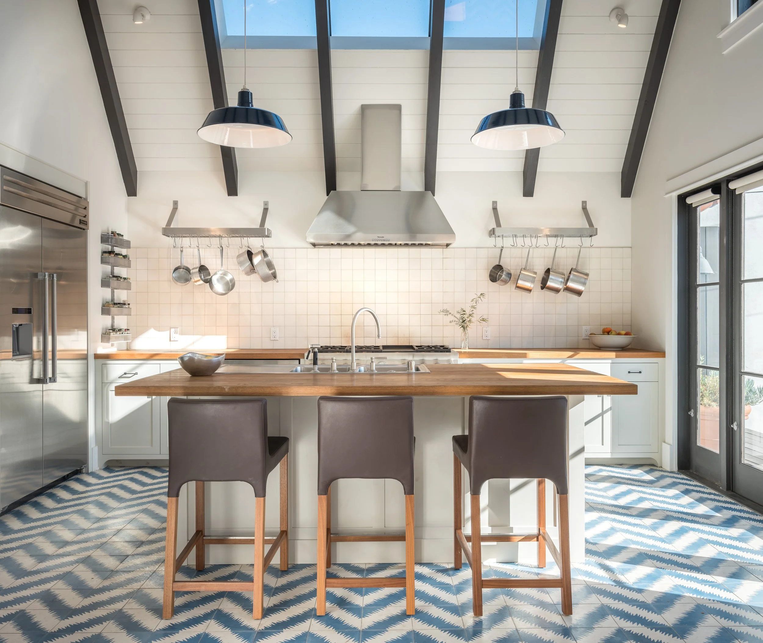 Modern kitchen with an island, barstools, hanging pots, skylights, and patterned tile floor.