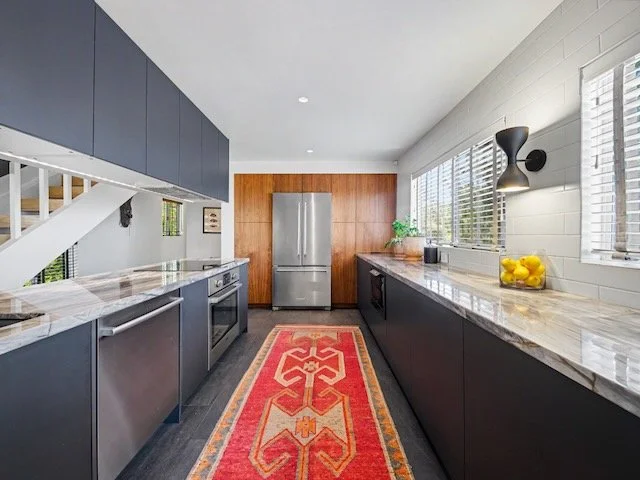 Modern kitchen with dark lower cabinets, light upper cabinets, marble countertops, stainless steel refrigerator, and a red patterned rug on dark flooring.