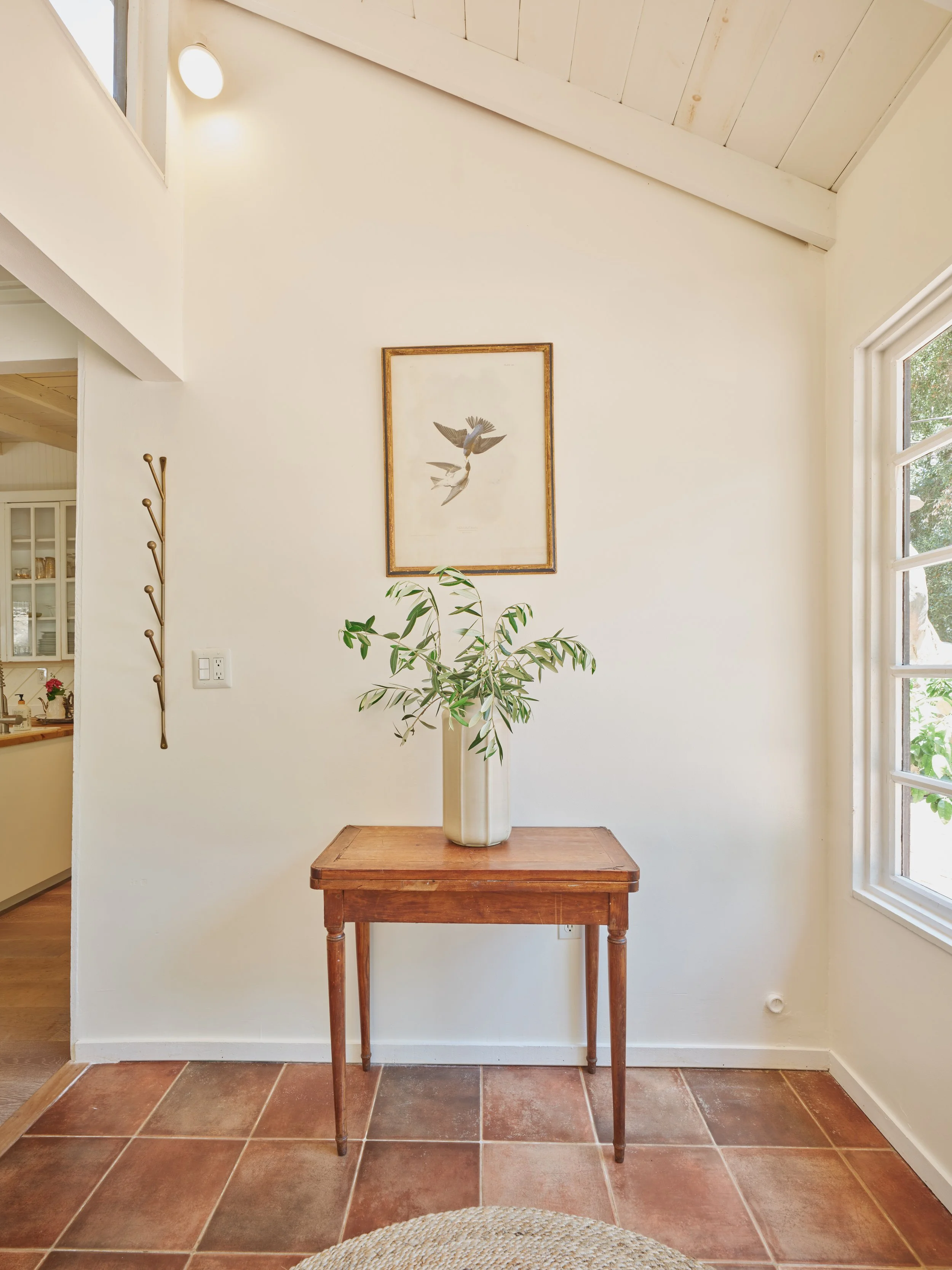 Interior entryway featuring a wooden table with a white vase holding leafy branches, a framed bird illustration on the wall, ceramic tiled floor, and a window allowing natural light.