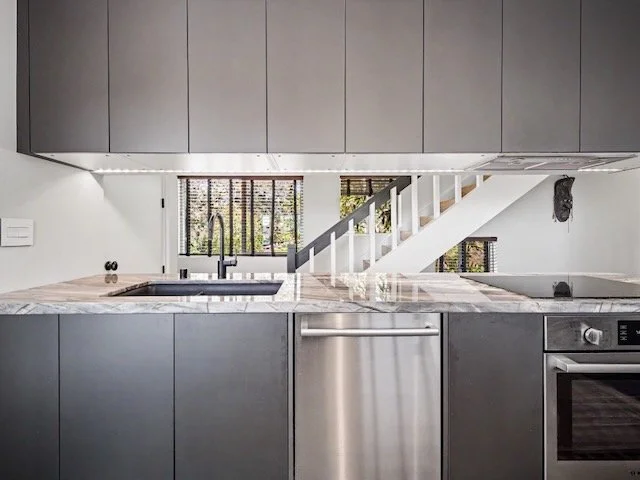 Modern kitchen with gray cabinetry, a marble countertop, stainless steel appliances, and a staircase leading upstairs.