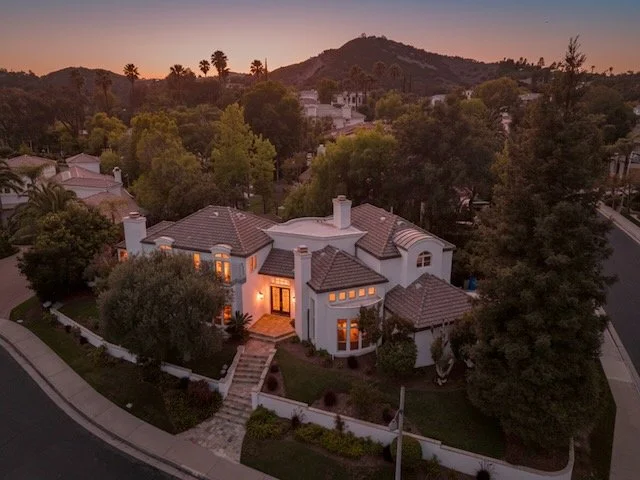 A large white house with a courtyard, surrounded by lush green trees and situated on a corner lot in a Calabasas neighborhood at dusk.