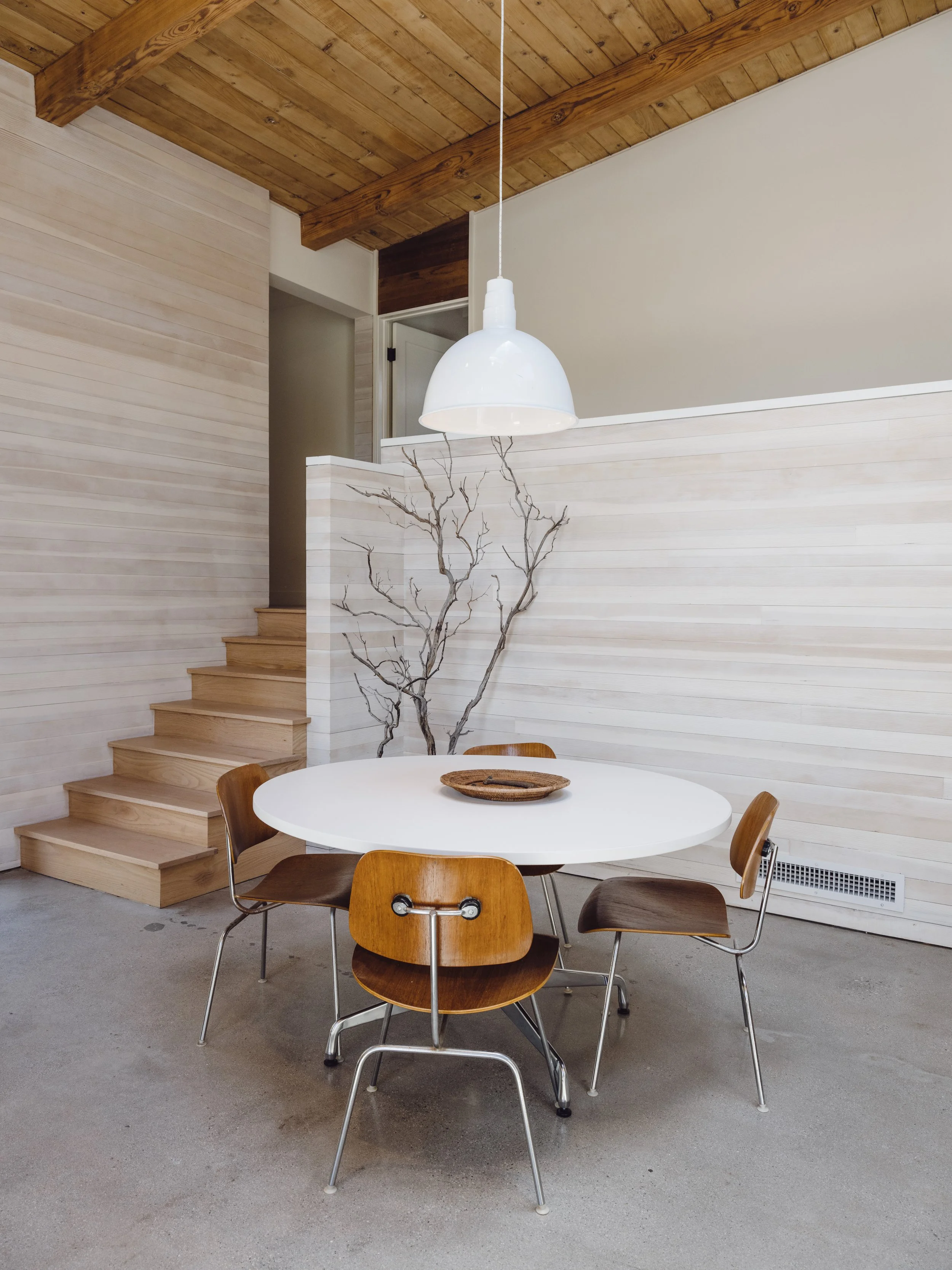 Modern dining area with a round white table, four wooden chairs, a white hanging lamp, wooden staircase, and decorative branch accents against wood-paneled walls and ceiling.