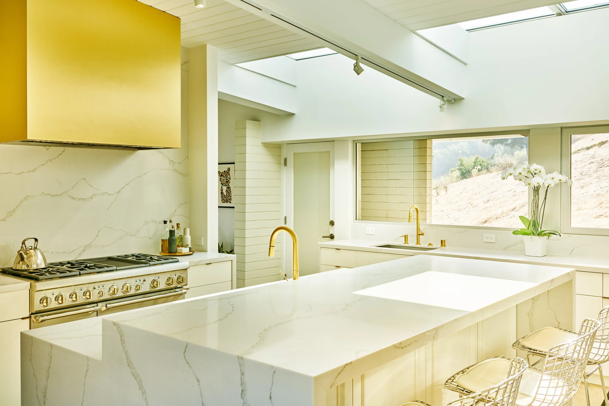 Modern kitchen with white marble countertops, gold fixtures, stainless steel stove, potted orchid on the counter, and large windows with scenic view.