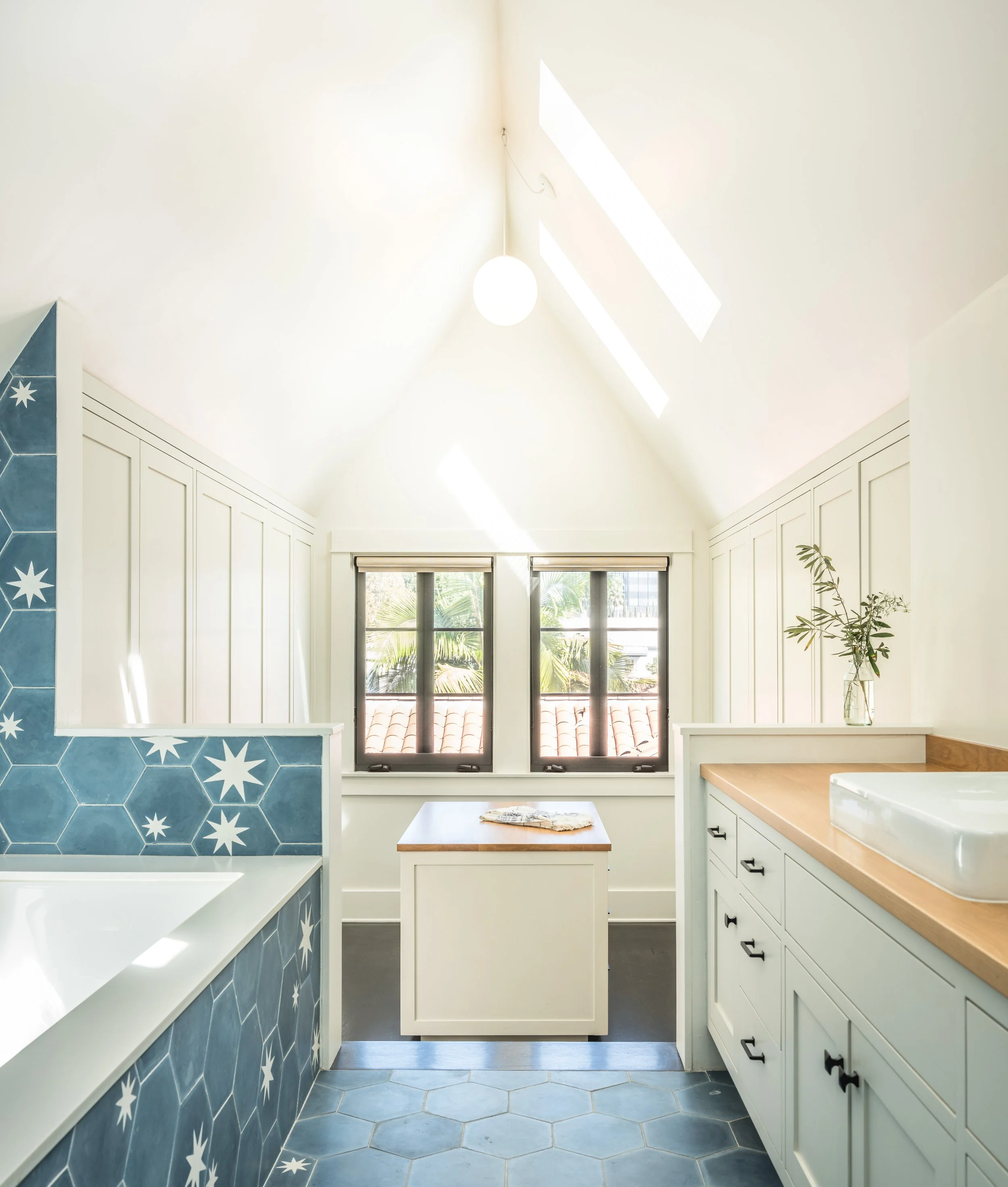 Bright modern bathroom with hexagonal blue tiles, white cabinets, a wooden countertop, a large bathtub, and a skylight. The room features an island in the center and a plant in a glass vase on the counter.