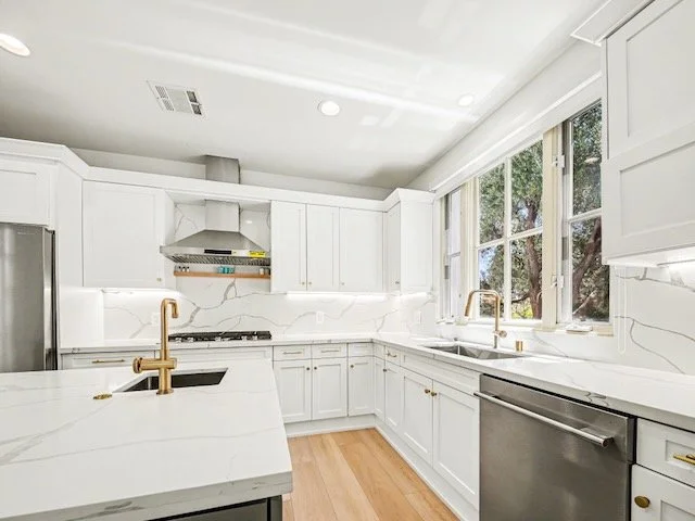 Modern kitchen with white cabinets, marble countertops, large windows, gold fixtures, stainless steel appliances, and hardwood floors.