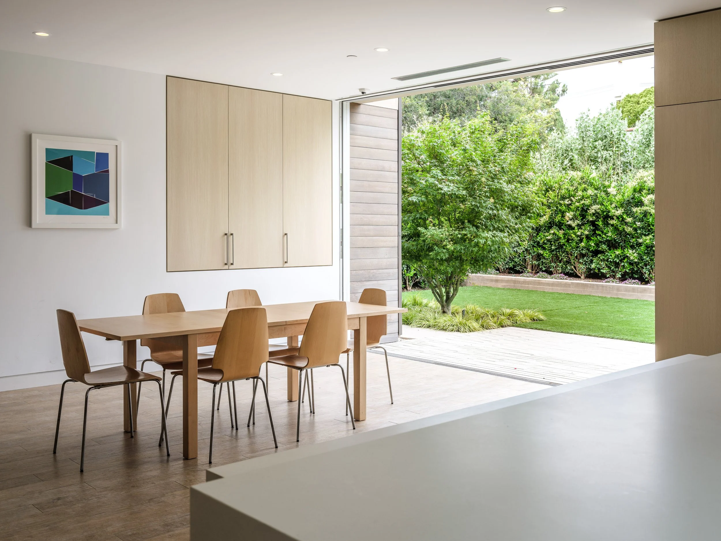 Modern dining room with wooden table and chairs, open sliding door to a green backyard, and geometric wall art.