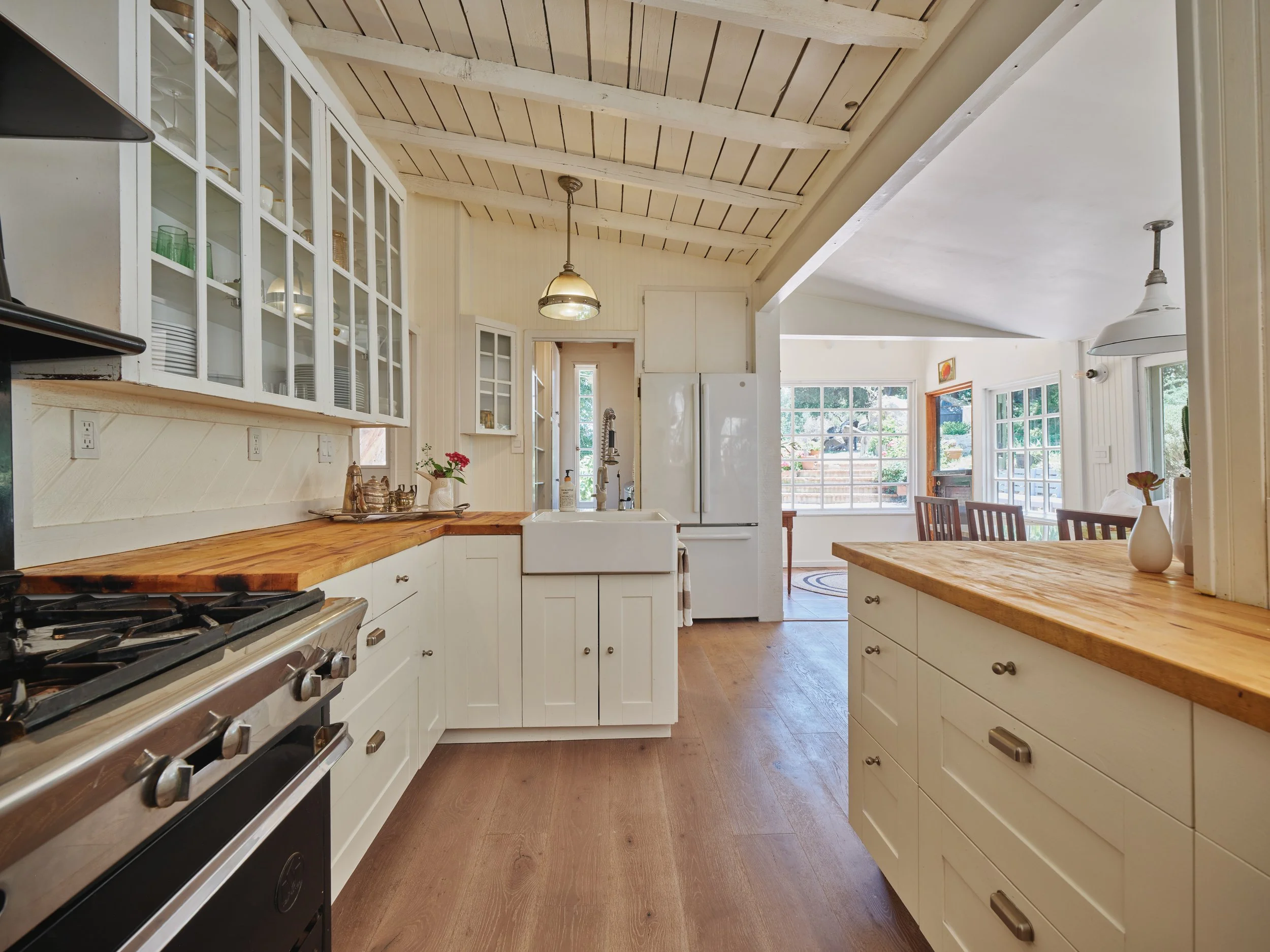 Spacious farmhouse-style kitchen with white cabinets, wooden countertops, a farmhouse sink, and a large stove. Glass-front cabinets display glassware and dishes. The kitchen opens to a dining area with wooden chairs and a view of large windows.