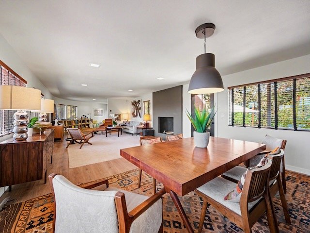 Spacious living and dining room with wooden furniture, a dining table with chairs, a sideboard, a cozy sitting area with armchairs, a large window with blinds, and a modern fireplace in the background.