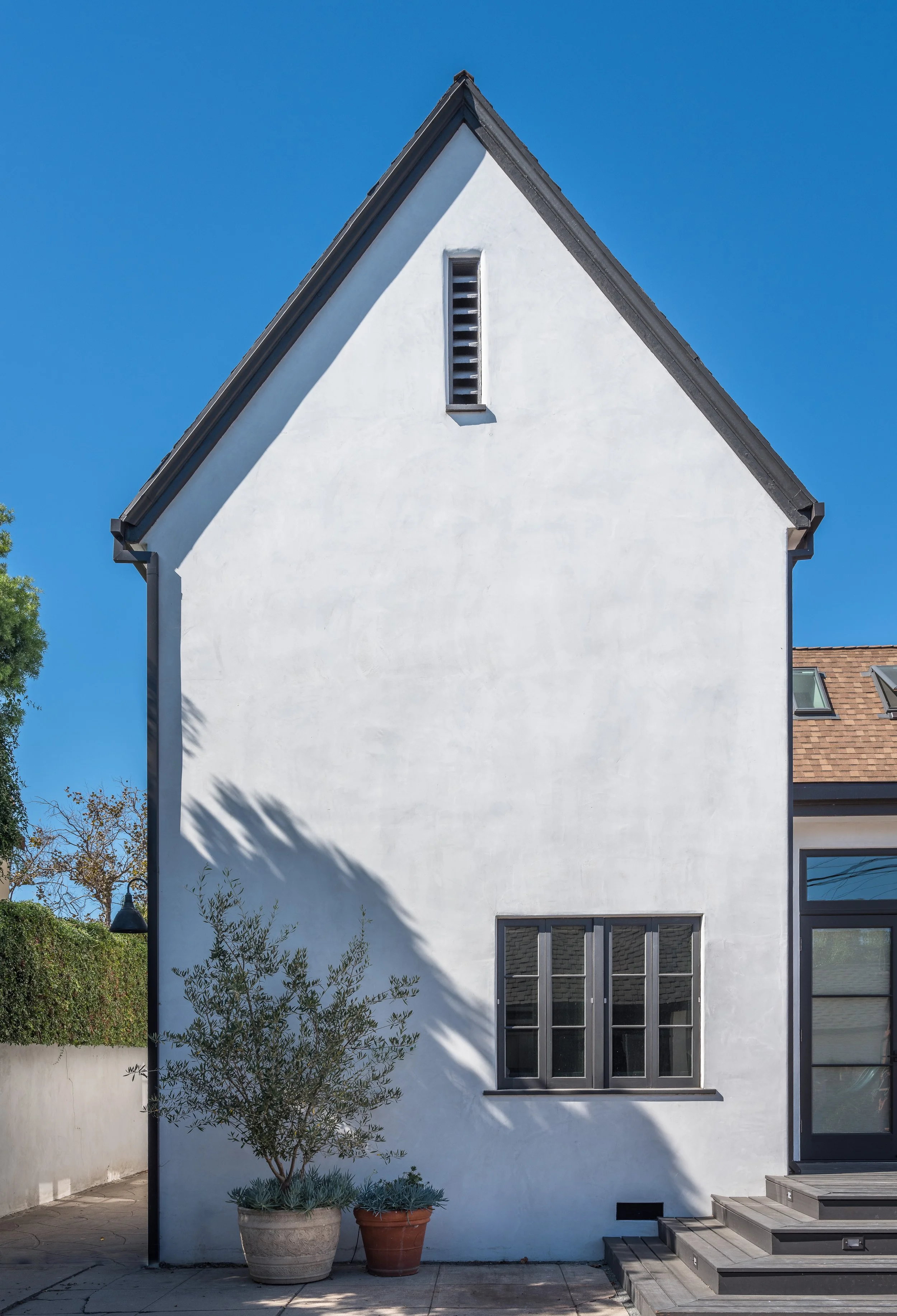 White stucco house exterior with a gable roof, small window, and vent. Two potted plants are placed near the wall next to concrete steps. The sky is clear and blue.