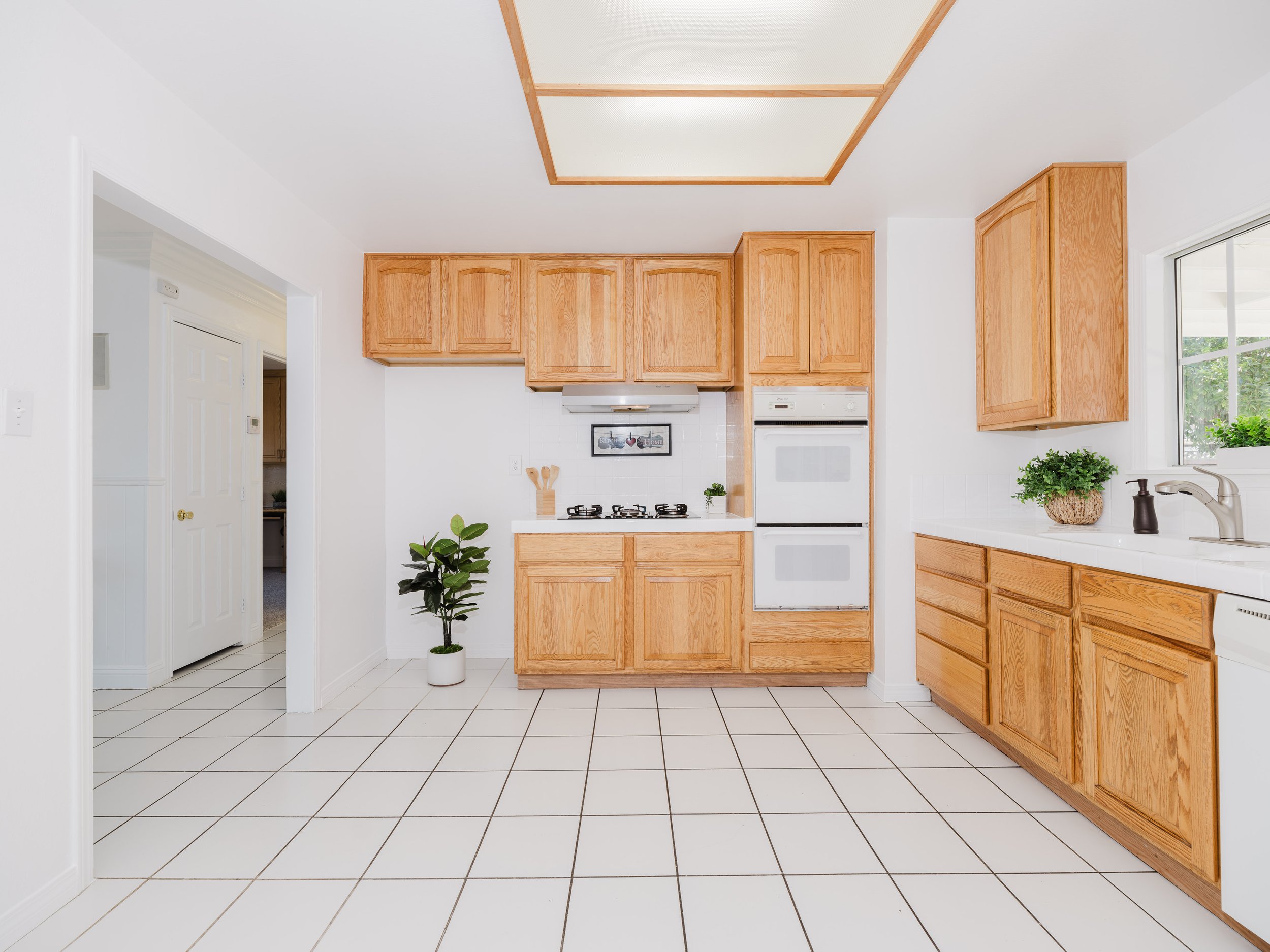 A modern kitchen with wooden cabinets, white tile flooring, built-in oven, stove, potted plants, and a window.