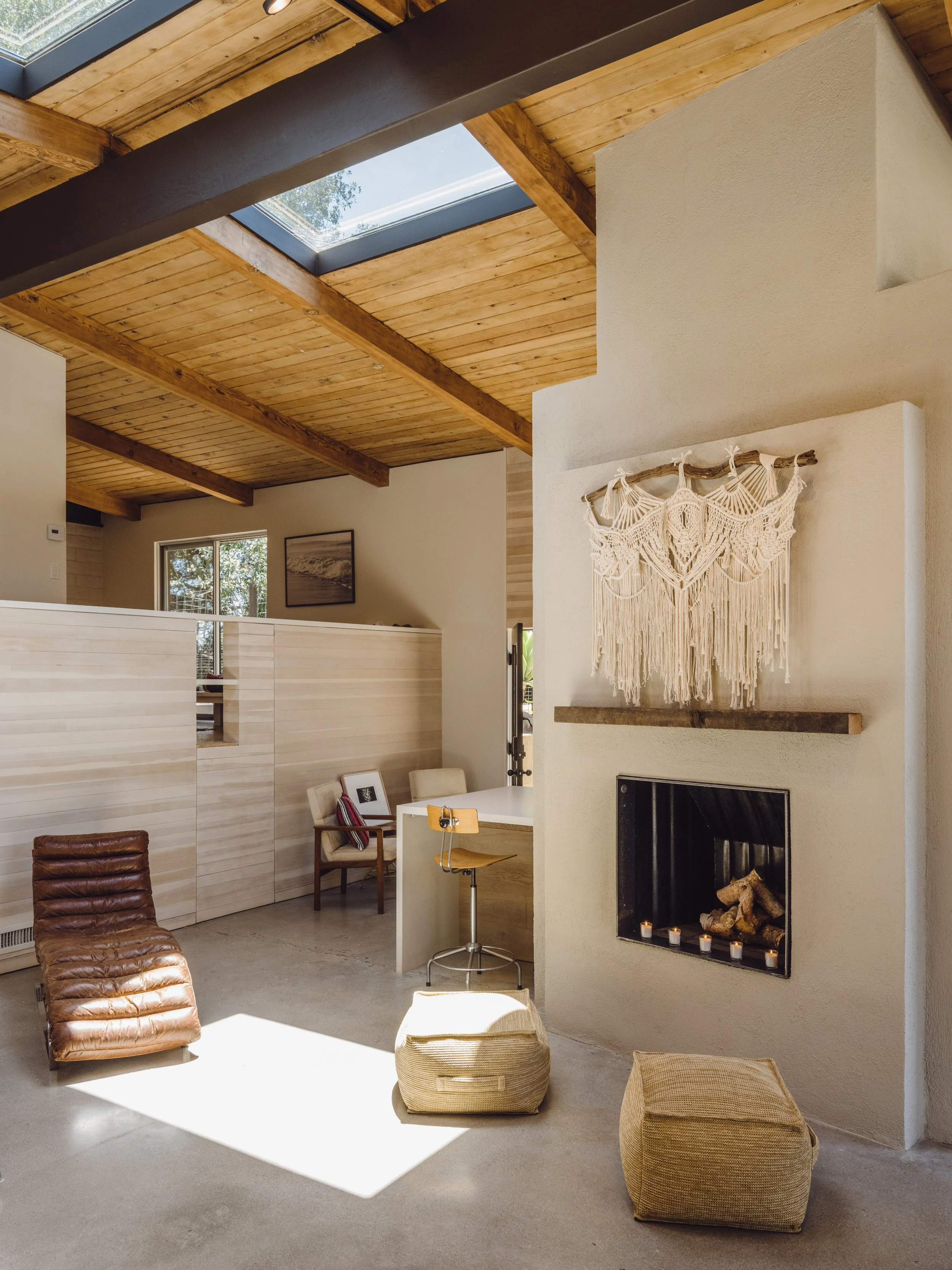 Modern living room with a wooden ceiling, skylight, and exposed beams. Features include a leather lounge chair, wooden chair, poufs, a built-in fireplace with candles, and a macrame wall hanging.