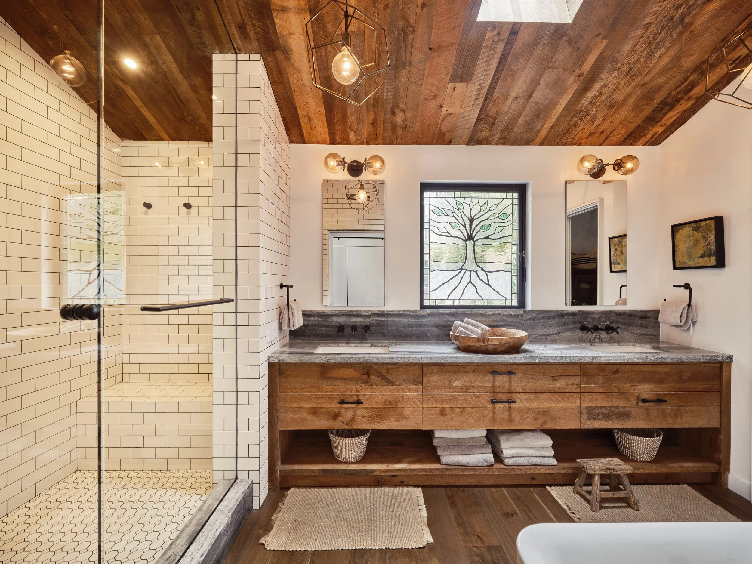 Rustic-style bathroom with a wooden ceiling, tiled shower area with glass door, large wooden vanity with dual sinks, dark stone countertop, and a stained glass window depicting a tree design.