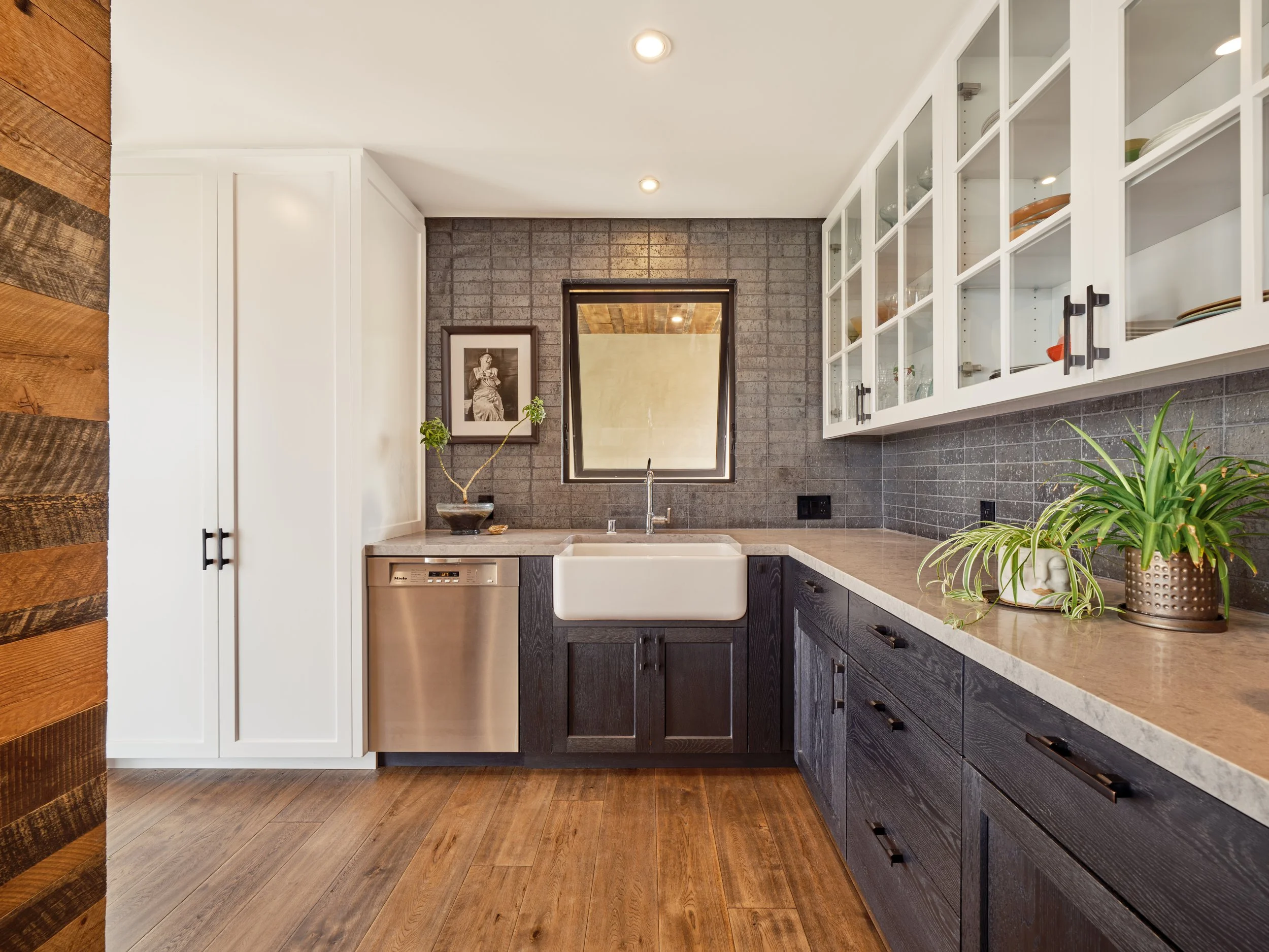Modern kitchen interior with wood flooring, farmhouse sink, black cabinets, gray tile backsplash, white upper cabinets with glass doors, stainless steel dishwasher, houseplants, and wall art.