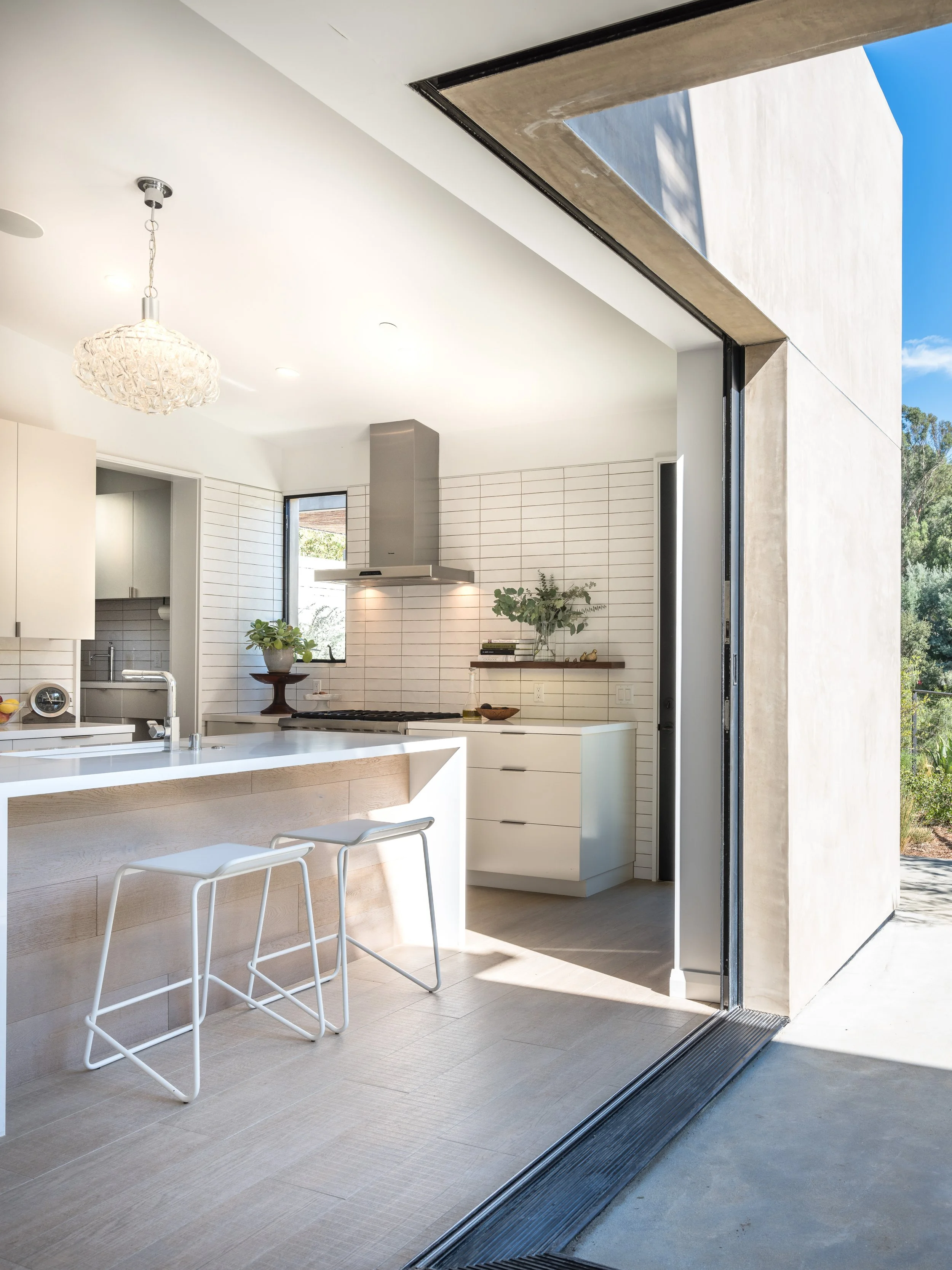 Modern kitchen with an open design, featuring minimalist white furnishings, bar stools, pendant light, and natural lighting from an open sliding door leading outside.