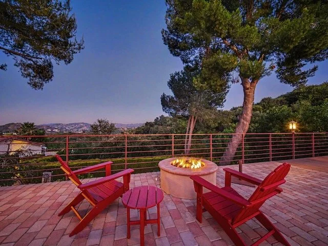 Outdoor patio with two red Adirondack chairs and a matching small side table, a fire pit with a fire, tall trees, a railing, and a scenic view of Topanga State Park in the distance at dusk.