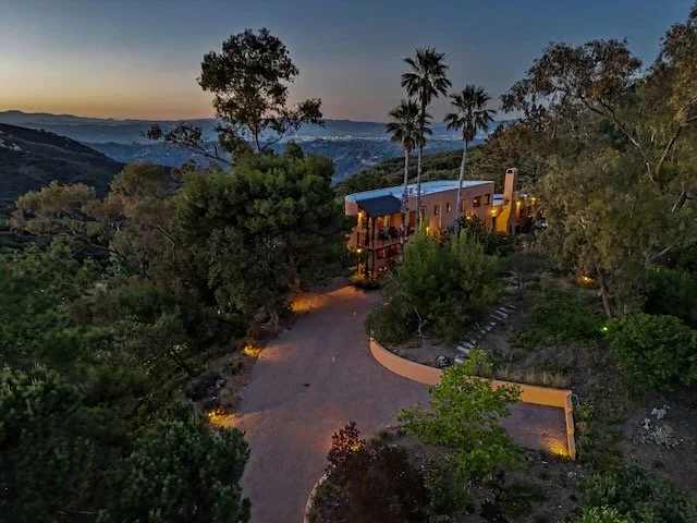 A house nestled among trees on a hillside at dusk, with a winding driveway leading to it and Topanga Canyon mountain views in the background.