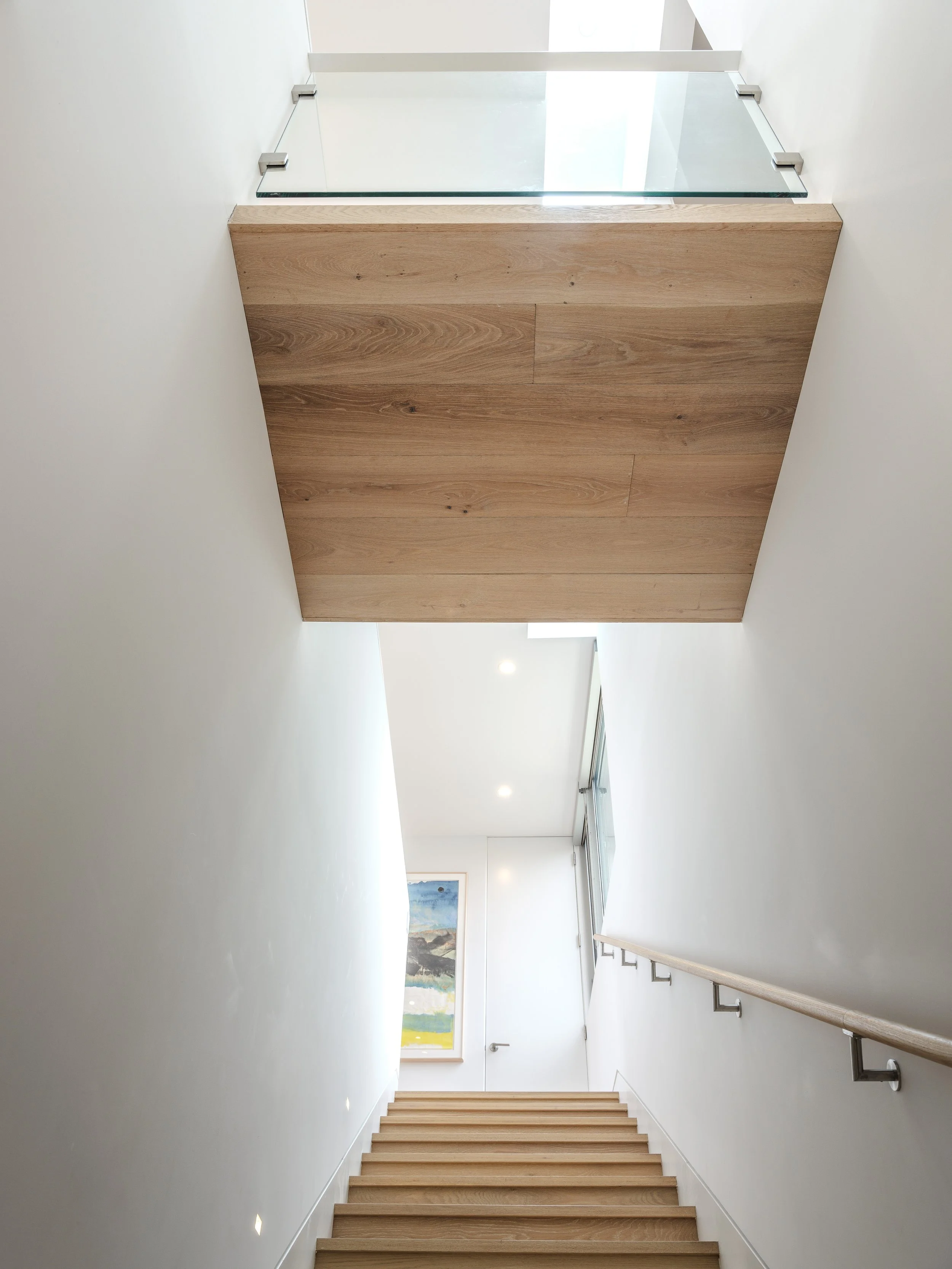 Modern indoor staircase with wooden steps leading to a well-lit hallway, featuring a framed landscape painting on the wall, wooden ceiling, and glass railing above.