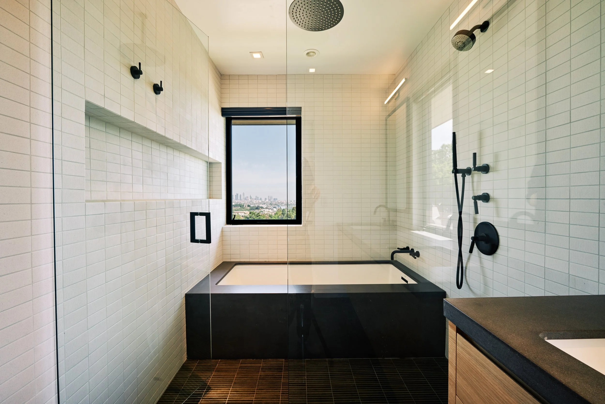 Modern bathroom with a glass-enclosed shower, black fixtures, a white tiled bathtub, and a large window with a city view.