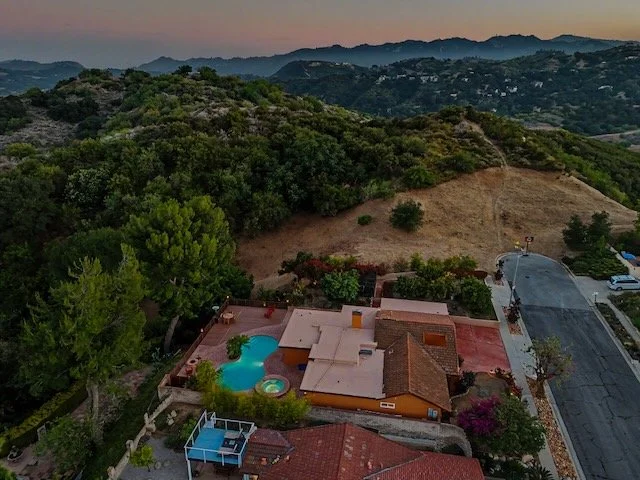 Aerial view of a residential house with a swimming pool, surrounded by trees and Topanga State Park in the background during sunset.