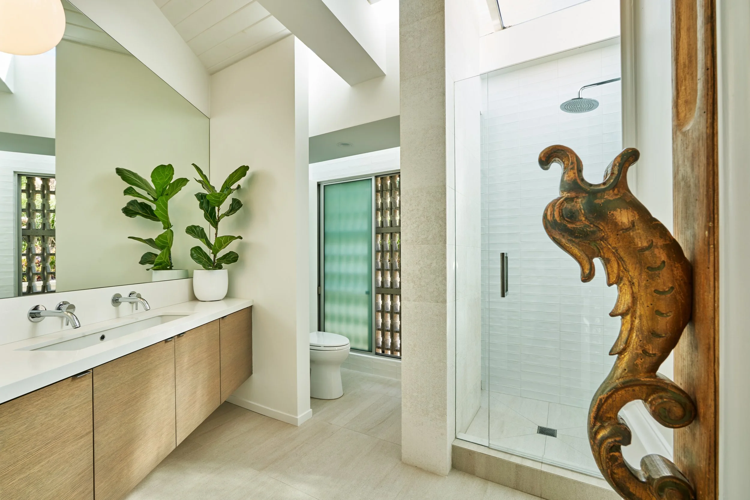 Modern bathroom with wood vanity, dual sinks, white countertop, potted plant, glass shower, and decorative wood accent.