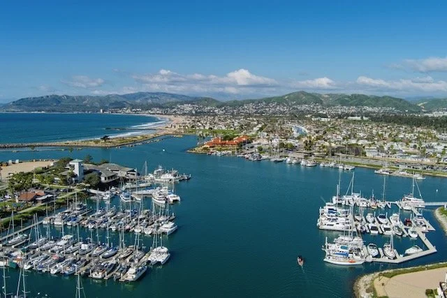 Aerial view of Oxnard marina with numerous sailboats and yachts docked, overlooking a coastal city with mountains in the background under a partly cloudy sky.