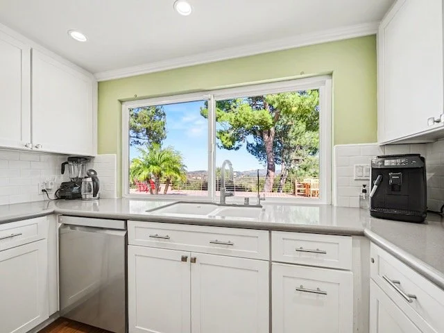 Kitchen with white cabinets, a large window overlooking trees and outdoors, a sink, a coffee machine, and small appliances on the counter.
