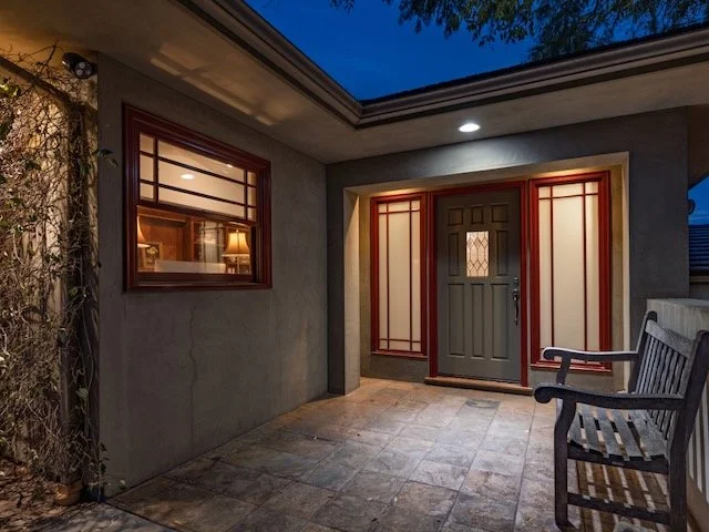 Front porch of a house during evening with a gray door, decorative red and beige trim, a window with a view inside showing lamps, a wooden bench, and a tiled floor.