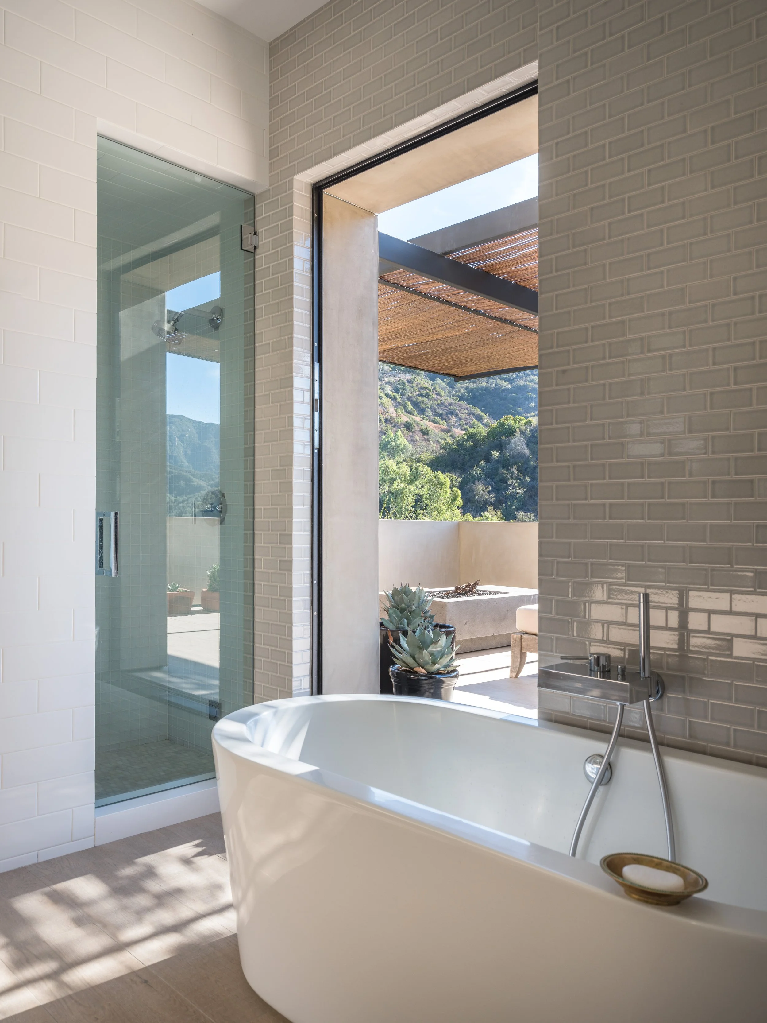 Modern bathroom with freestanding bathtub, glass shower enclosure, tiled walls, and outdoor view of mountains and plants.