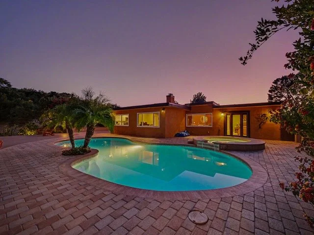 A backyard at dusk with a lit swimming pool, a single-story house with large windows, and a landscape of trees and hills in the background.
