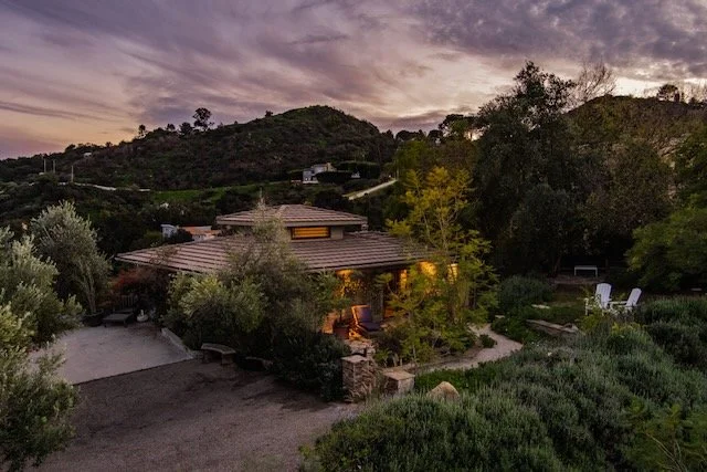 Landscape view of a house surrounded by trees and bushes at dusk, with hills and a cloudy sky in the background.