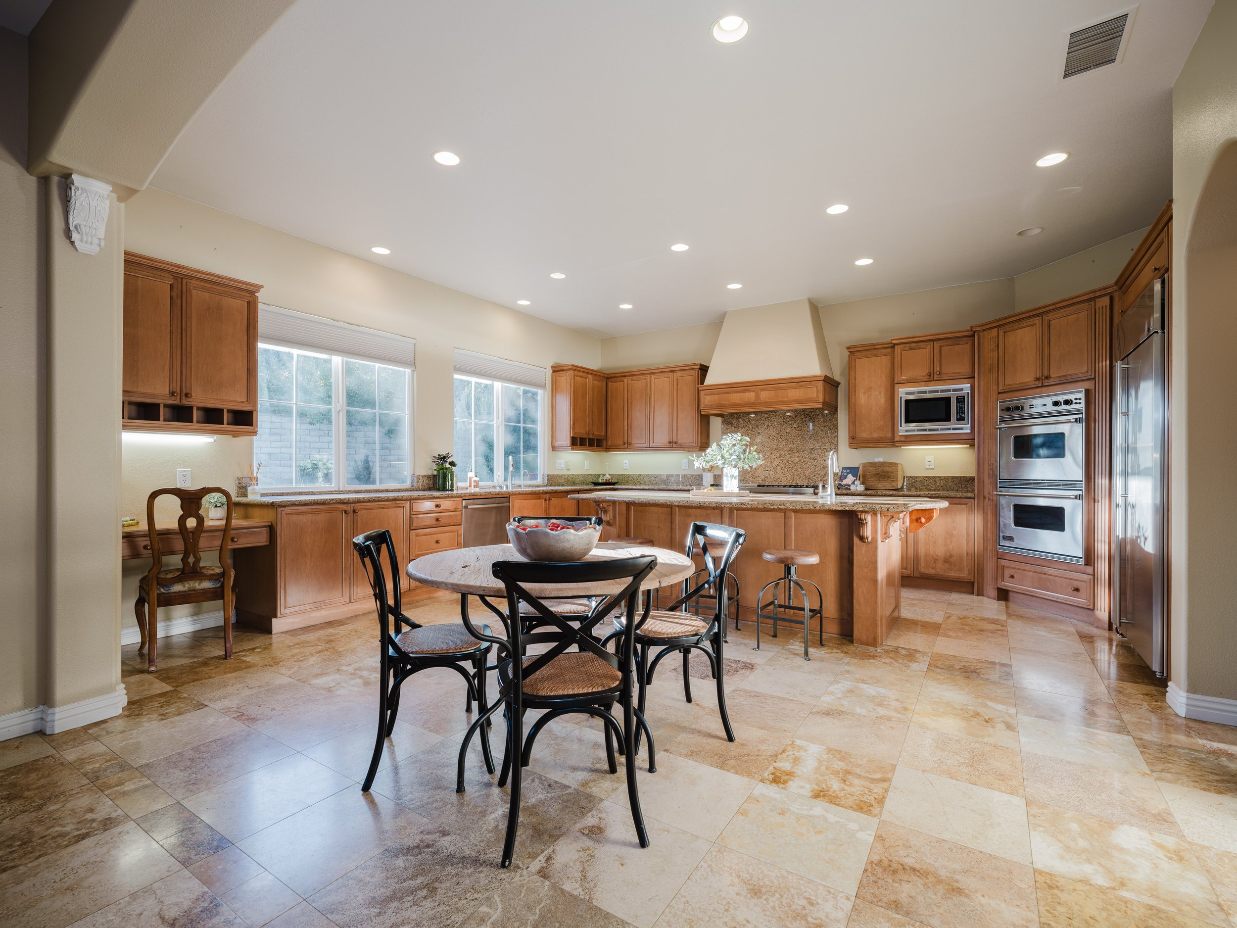 Spacious kitchen interior with wooden cabinets, stone tile flooring, and a central round table with chairs. Features include stainless steel appliances, a kitchen island, and large windows.