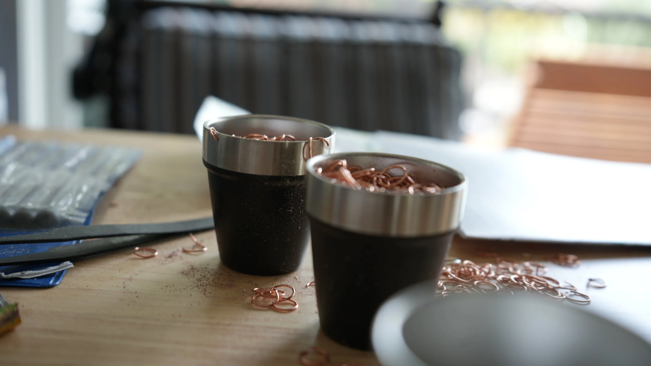 Close-up of two black coffee cups with metallic rims, filled with copper-colored metal shavings, on a wooden table with some spilled shavings and a blurred background.