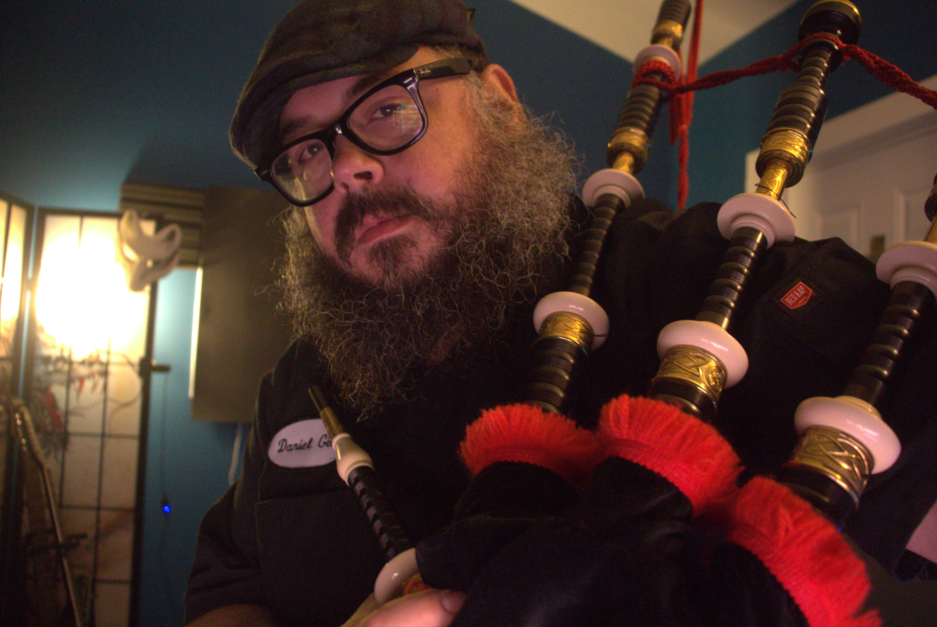 A man with a beard and glasses wearing a hat, playing a set of bagpipes in a room with blue walls, a guitar in the background, and a bright light source on the left side.