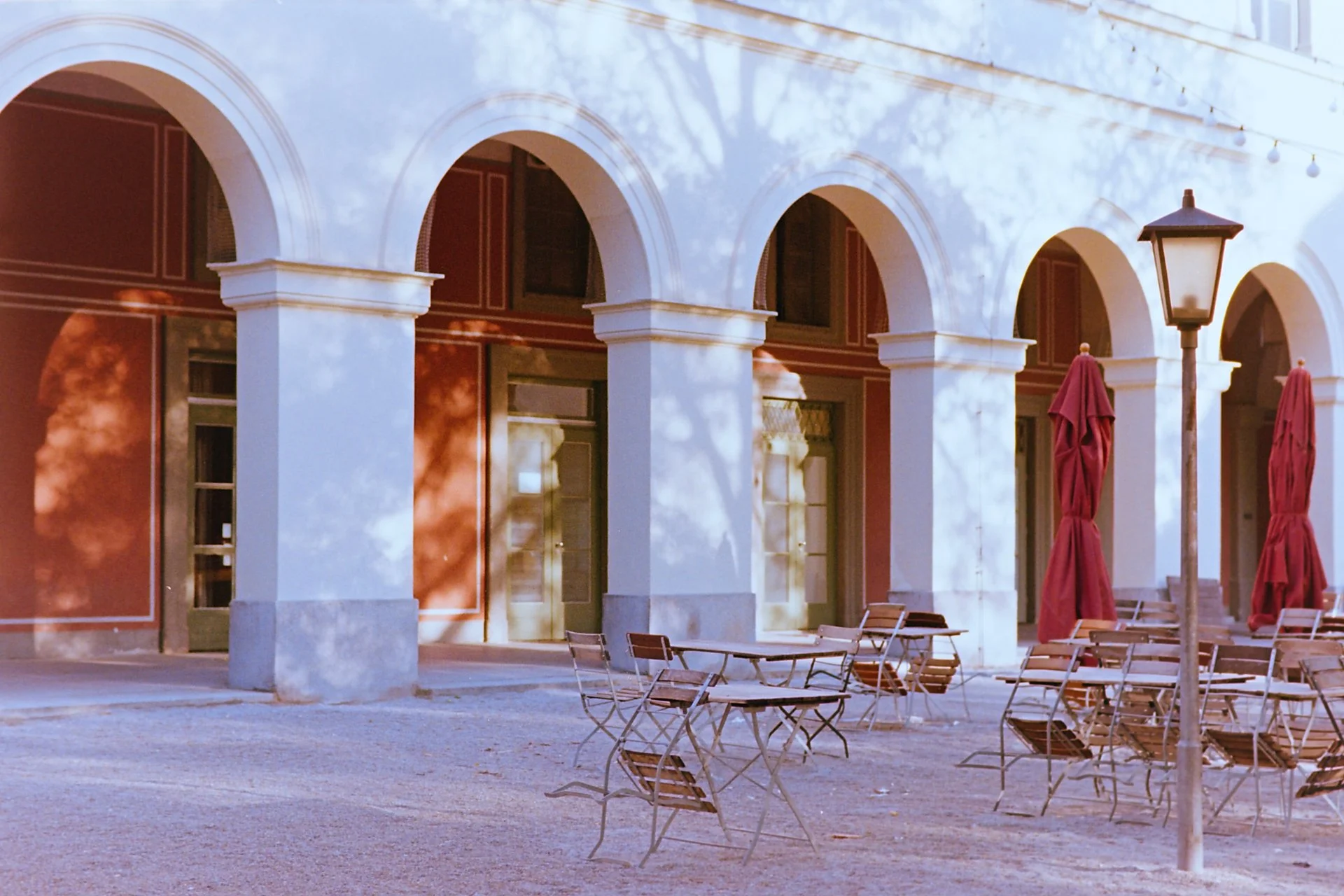 Hofgarten arcades behind Schuhmann’s Bar with empty tables, red parasols, and sunlit courtyard, shot on 35mm film.