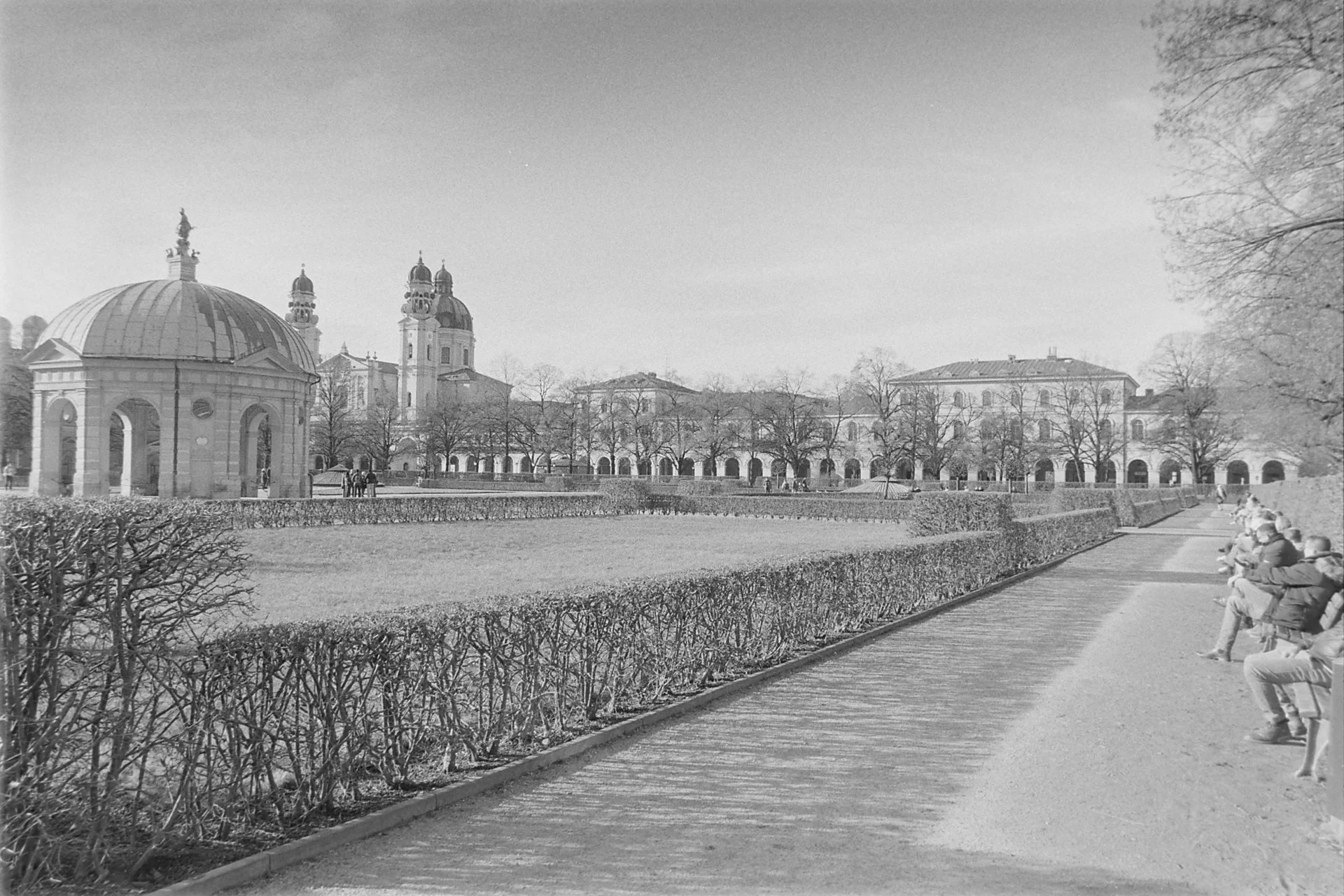 Black‑and‑white 35mm film photograph of Munich Hofgarten with Dianatempel, Theatinerkirche, and people sitting in winter sunlight