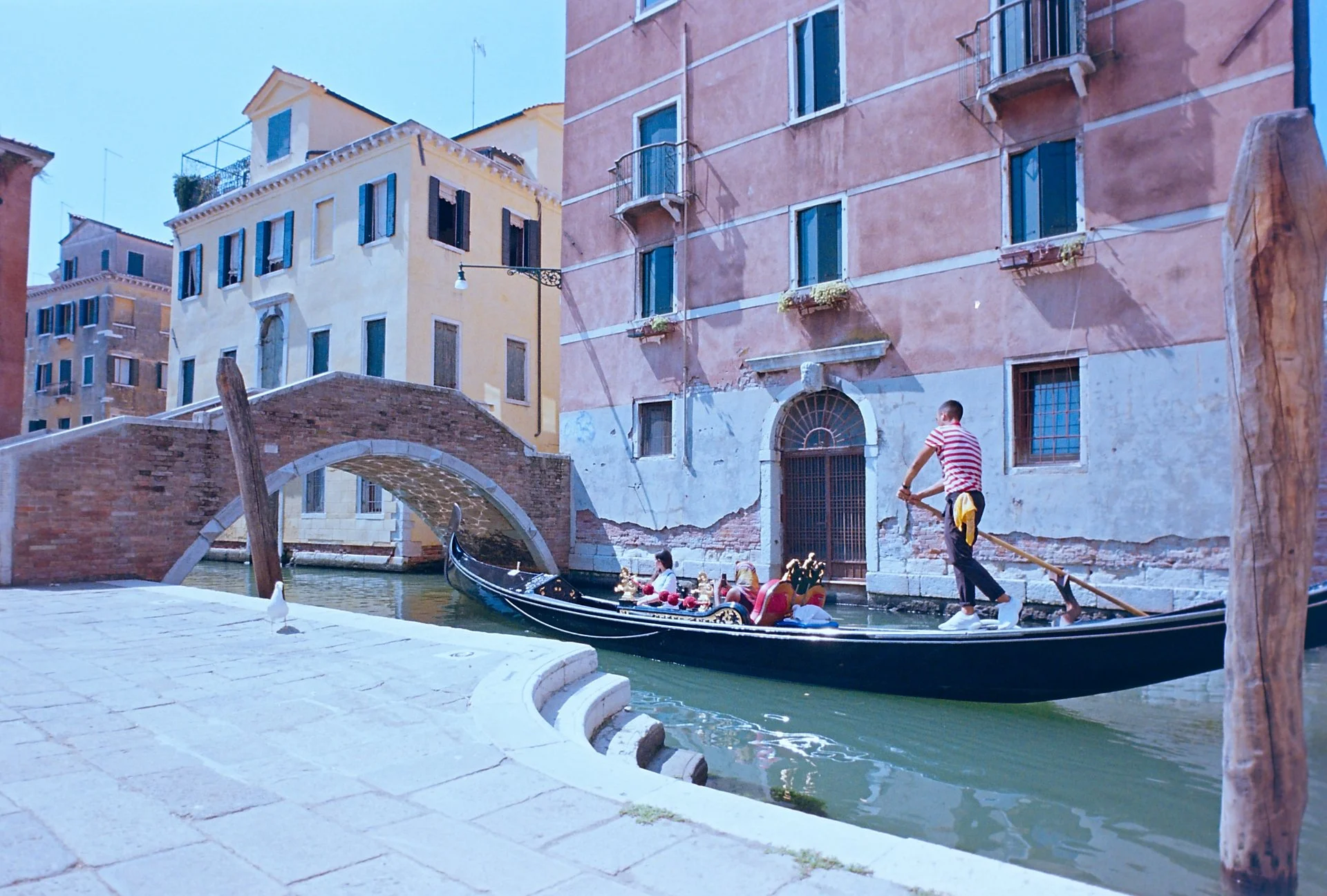 Gondola in a small Venetian canal approaching a bridge, with red and yellow buildings and stone steps, shot on 35mm film.