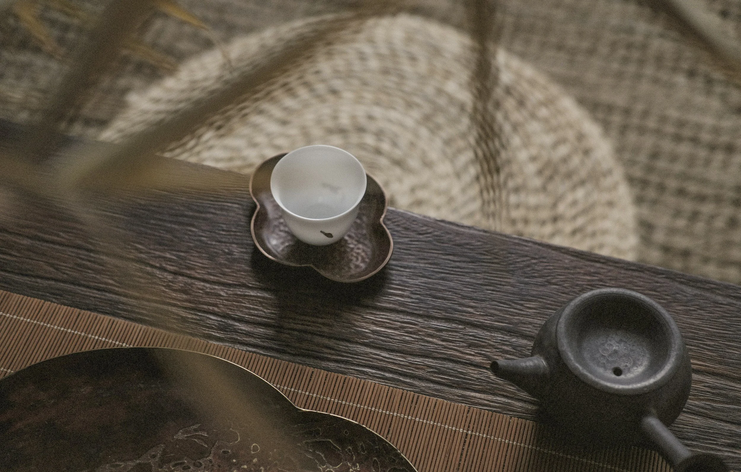 Top-down view of a wooden table with a small white cup on a black flower-shaped saucer and a black teapot nearby, with a woven placemat partially visible.