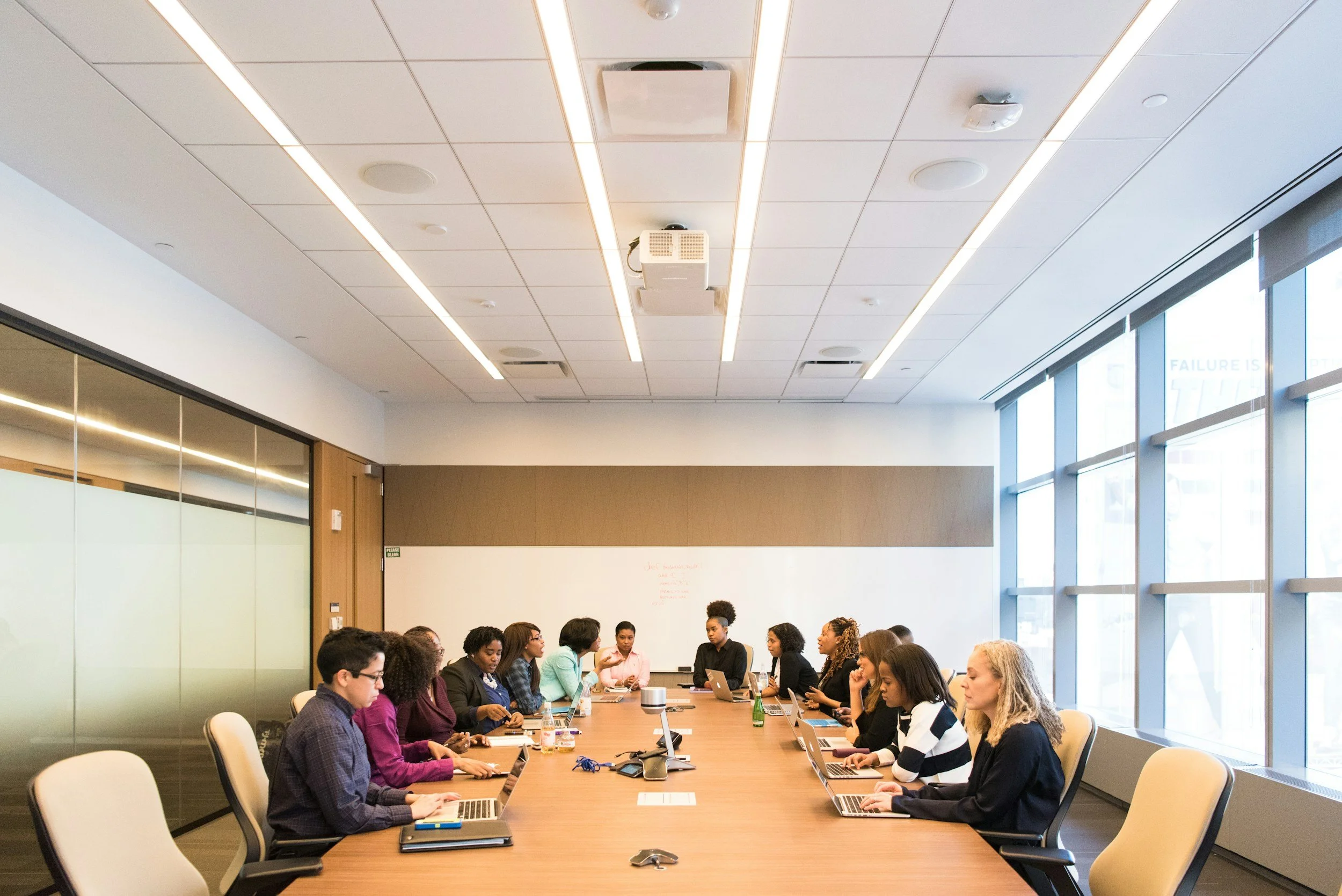 A diverse group of people sitting around a long conference table in a modern office meeting room, working on laptops and engaged in discussion.
