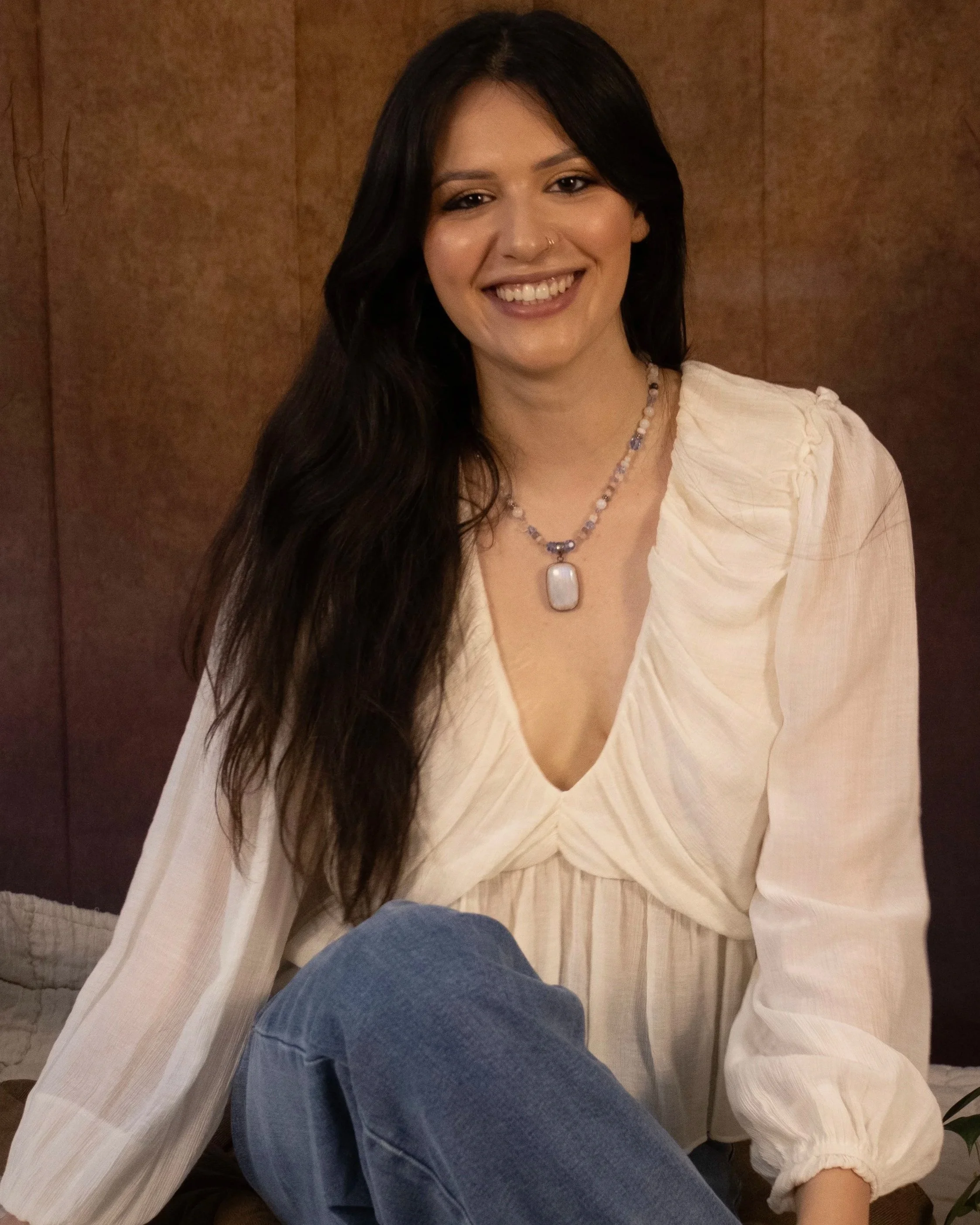 A woman with long dark hair, wearing a white blouse with puffed sleeves and a pearl necklace with a large pendant, smiles at the camera against a brown wooden background.